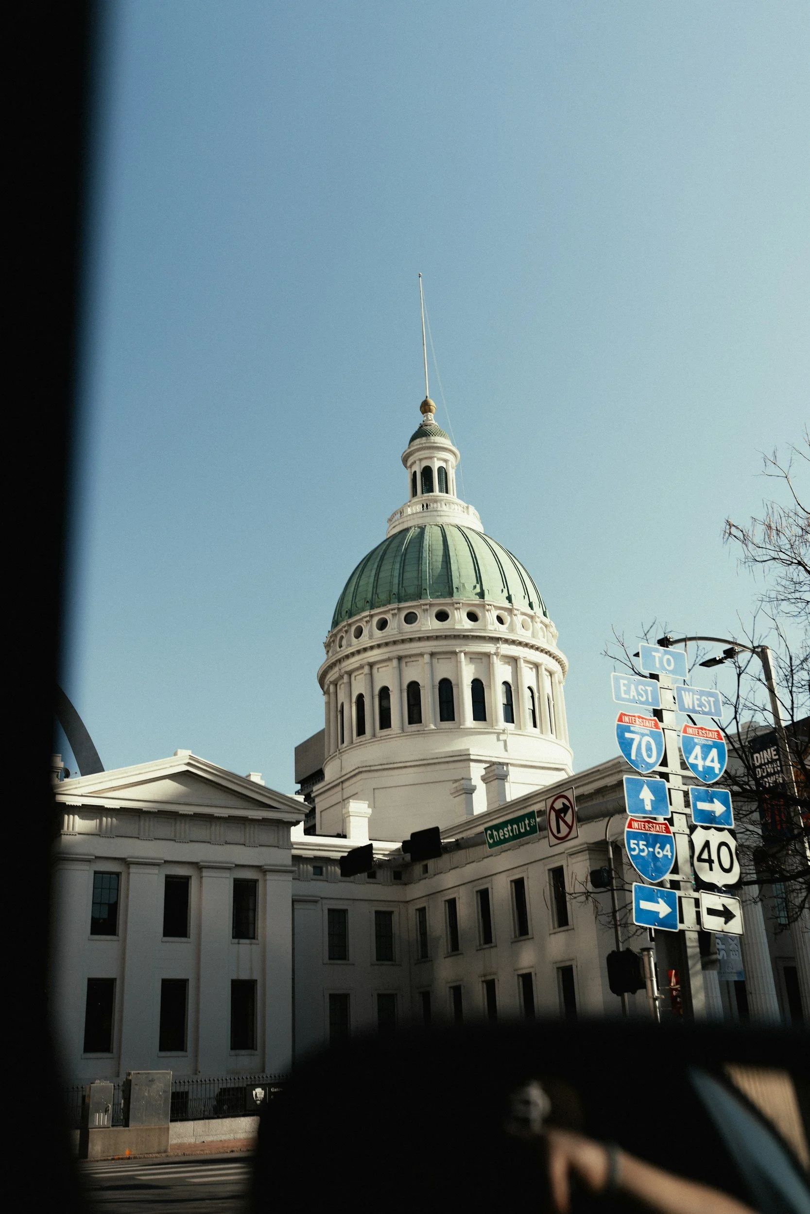 View of the California State Capitol building, with its distinctive dome and a clear blue sky in the background. Street signs are visible in the foreground, indicating directions and highways.
