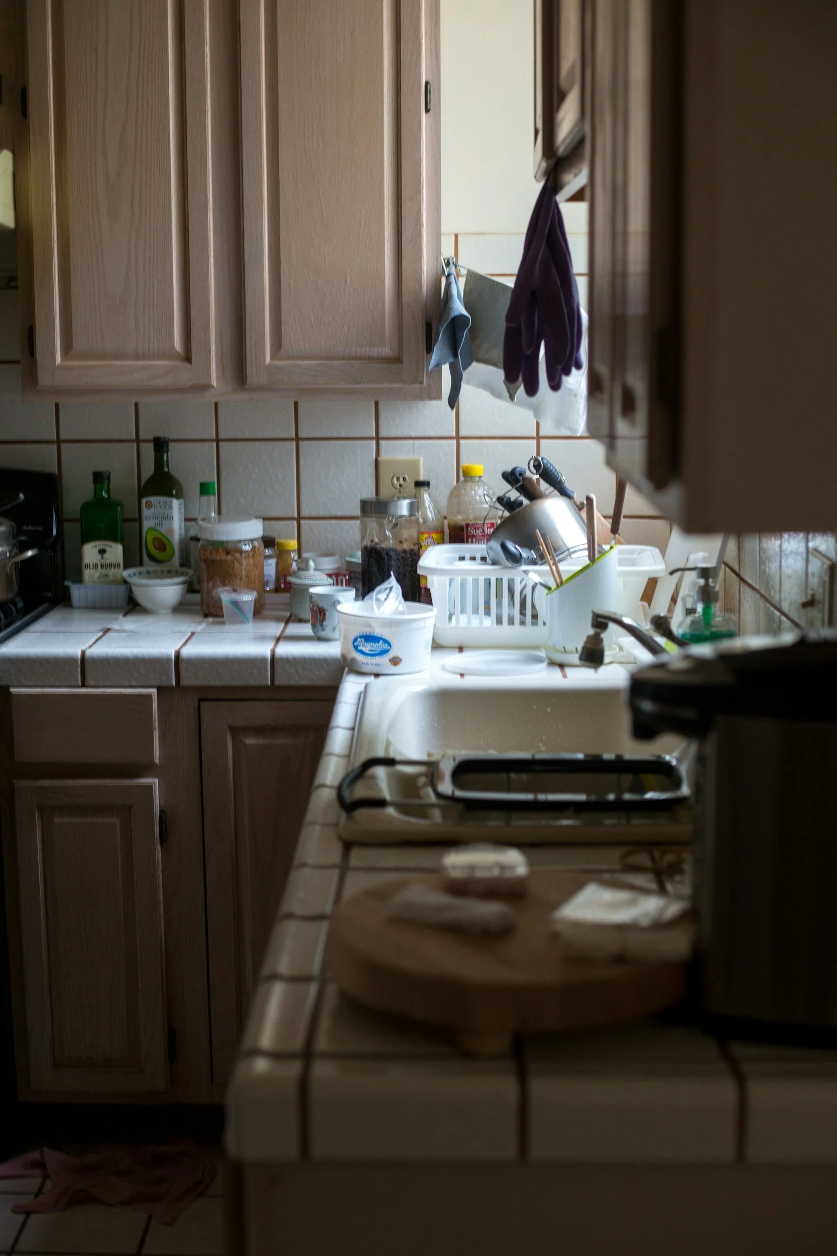 Cluttered kitchen countertop with various bottles, jars, dish soap, and a white dish drainer. Wooden cabinets above and a stove to the left. Haphazardly hung dish towels and gloves on a rack. Part of the sink and a towel on the floor are visible.
