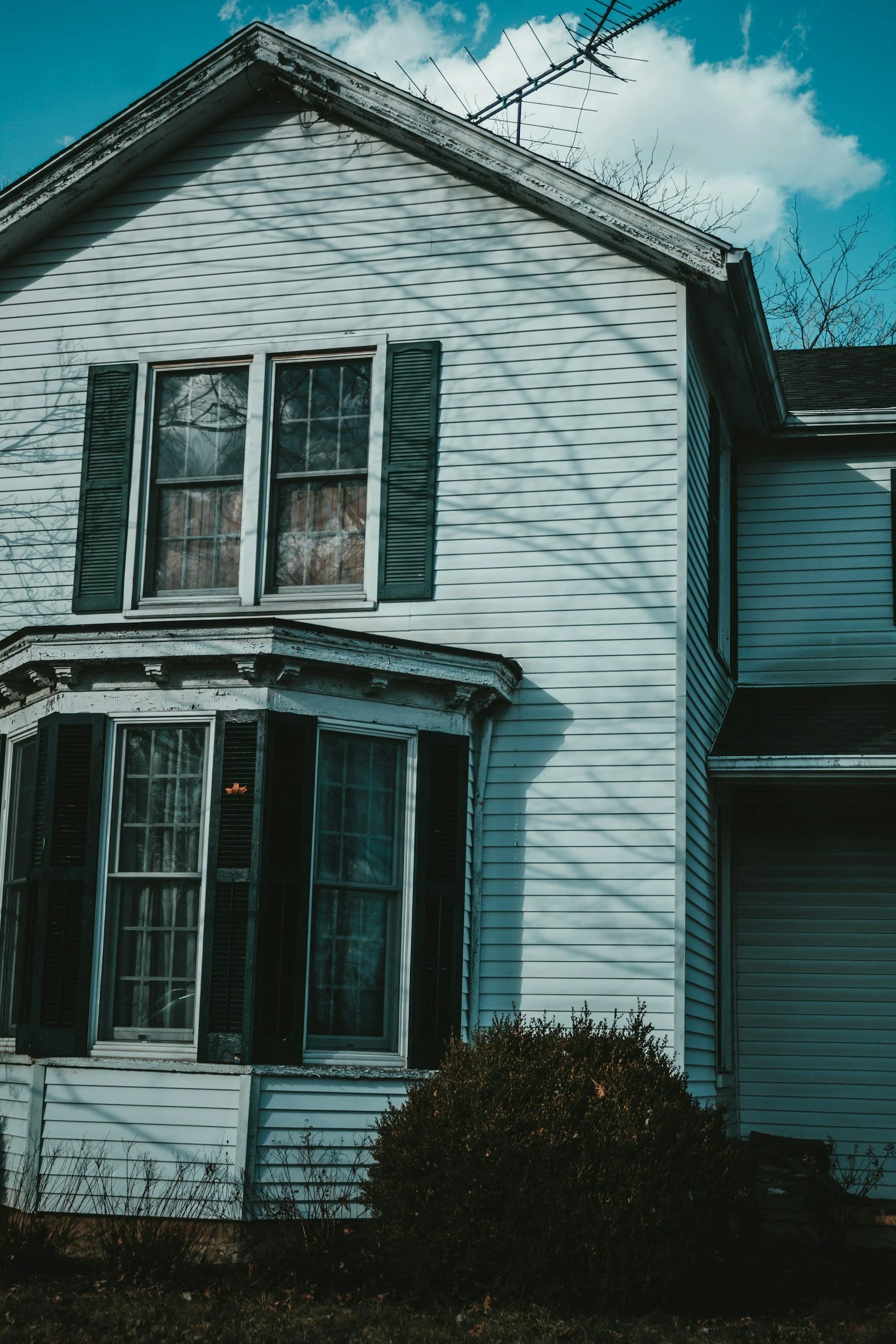 A side view of a white house with black shutters and multiple stories. The house has a gabled roof with an TV antenna on top. Shadows of tree branches are cast across the house's facade. There are bare trees and bushes in the foreground, under a partly cloudy sky.