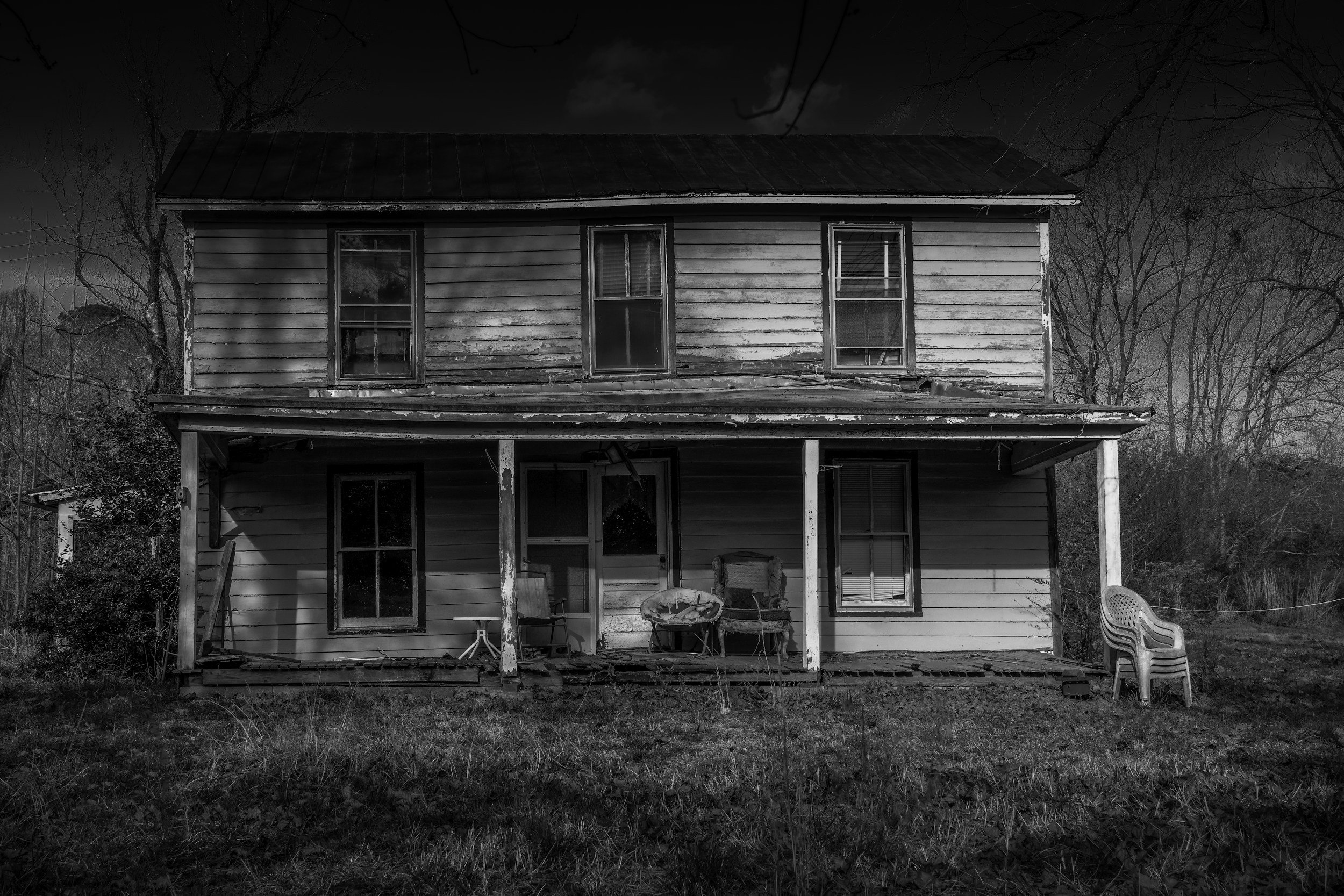 An old, dilapidated two-story house with a porch, missing some support beams, surrounded by leafless trees, and outdoor furniture including chairs, in black and white.