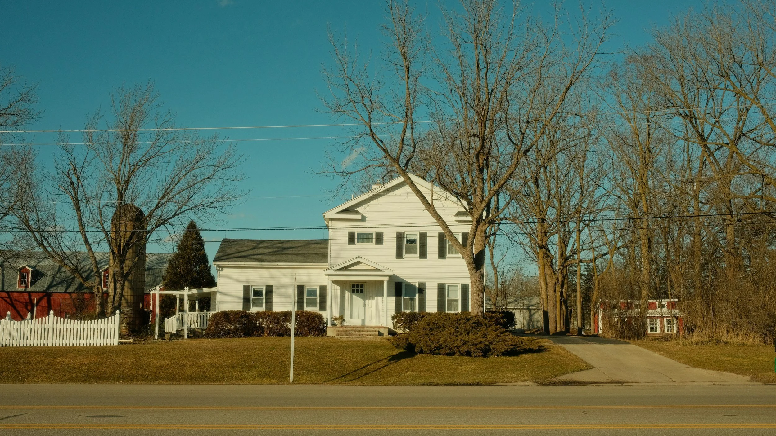 A two-story white house with a front porch and multiple windows, surrounded by trees with no leaves, a white picket fence, and a neatly trimmed lawn.