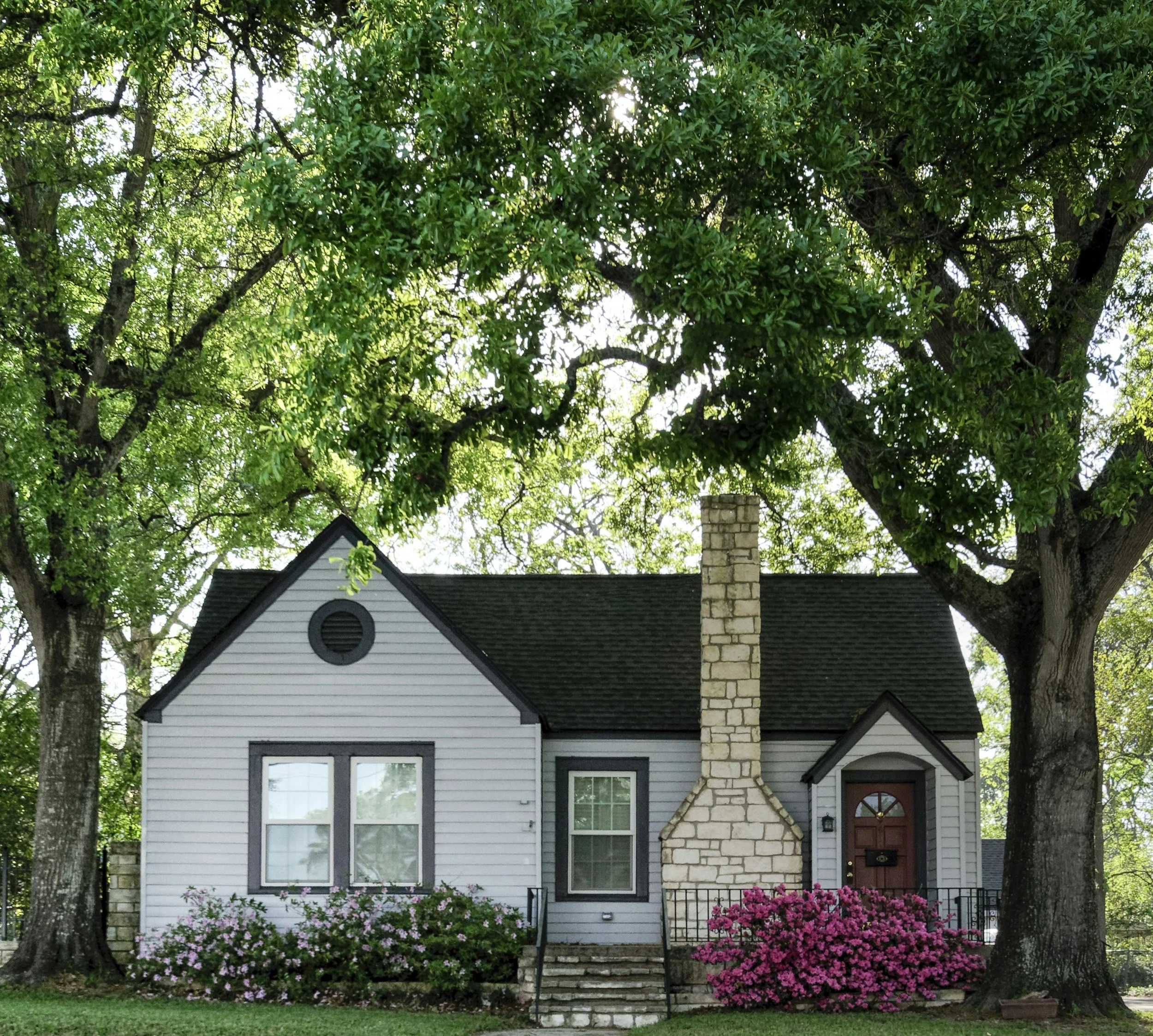A house with gray siding and black roof, surrounded by trees and pink flowers, with a brick chimney.