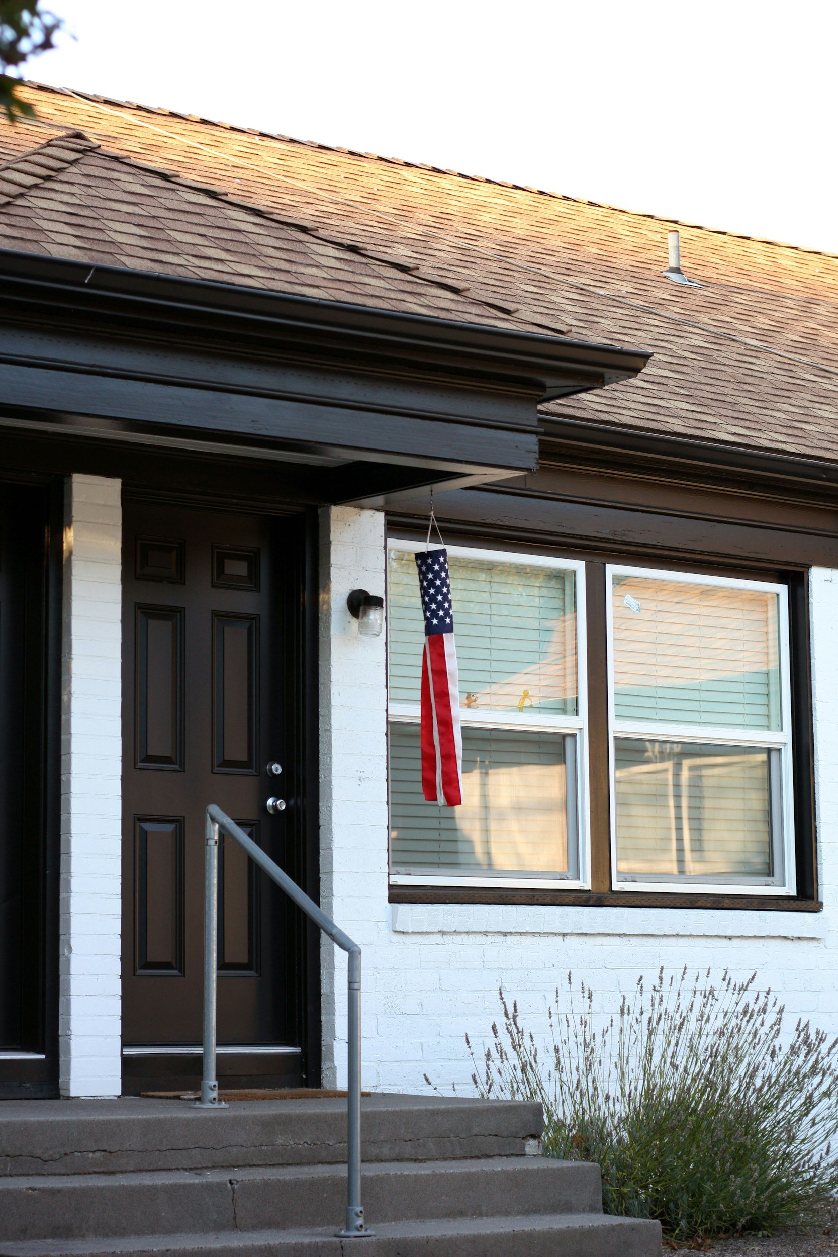 Front porch of a house with steps, black door, large window, and an American flag hanging beside the door.