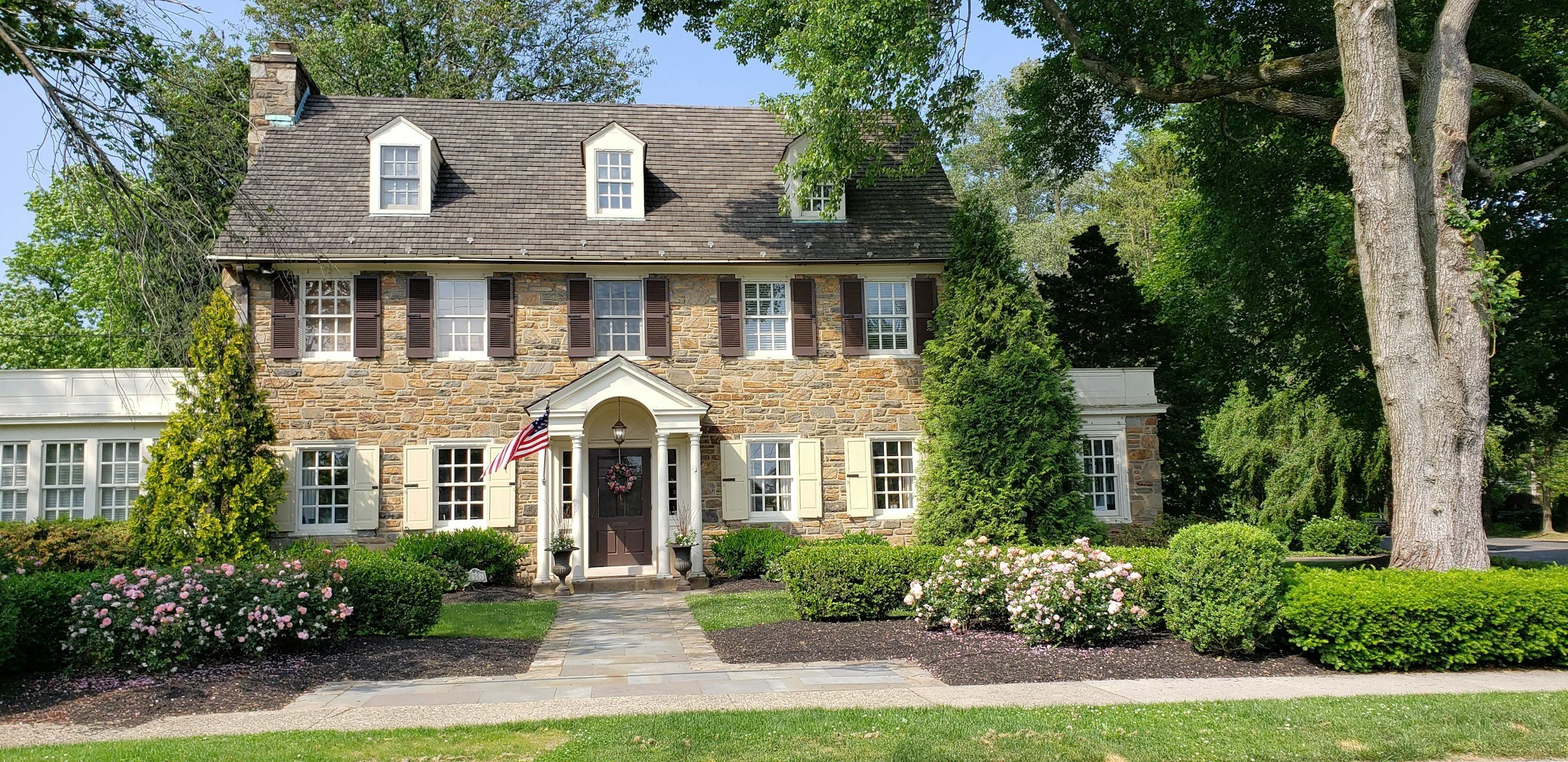 A large house with a stone facade, white window shutters, and dormer windows on the roof. The house has a small front porch with an American flag, potted plants, and a wreath on the door. The front yard has flowers, bushes, and trees, with a flagstone pathway leading to the entrance.