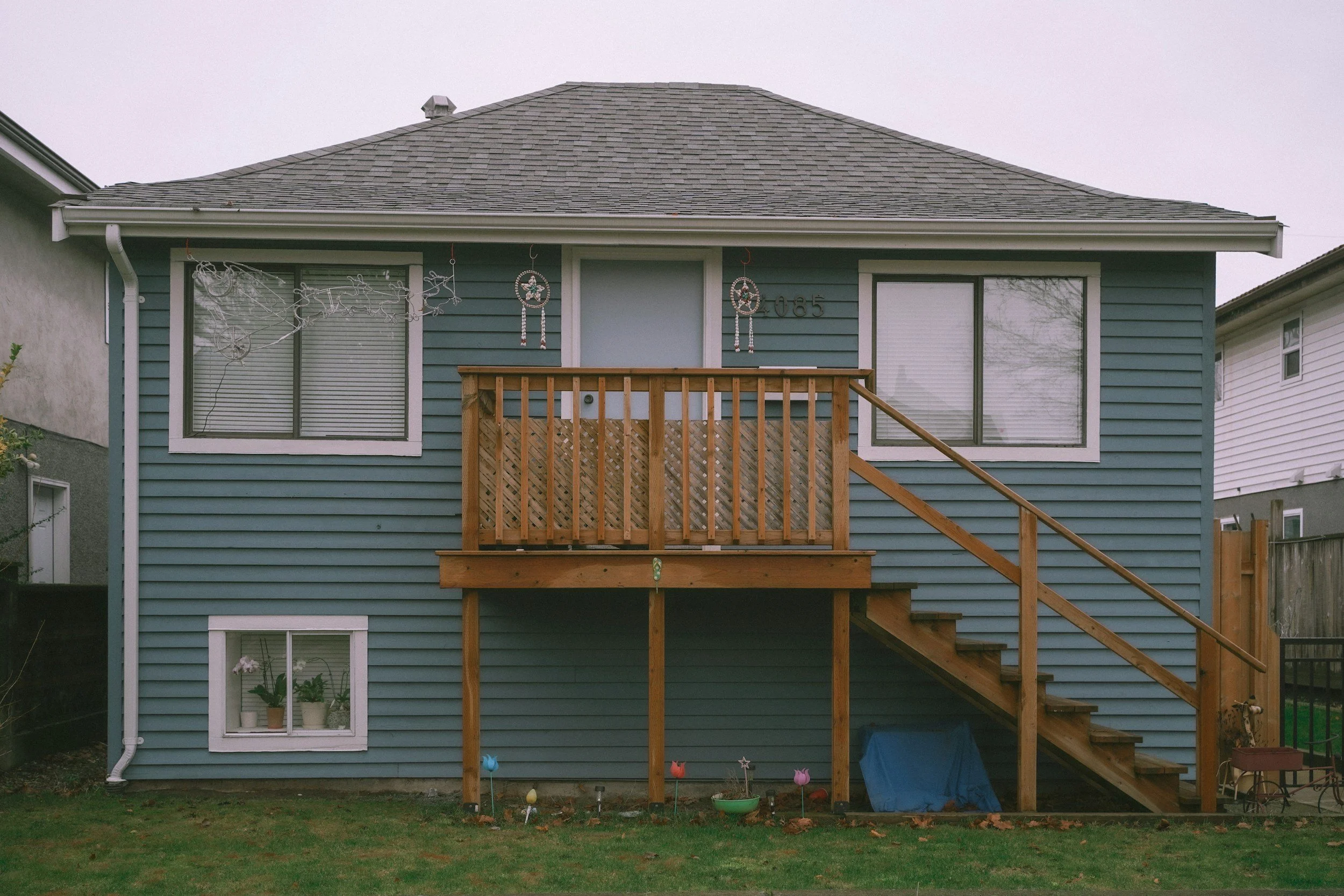 Back of a blue house with a small wooden deck and staircase. The house has two large windows with closed blinds, and holiday decorations hang in front of the upper window. There are potted plants on the lower window sill, and a garden area with decorative stakes and a small blue slide beneath the deck.