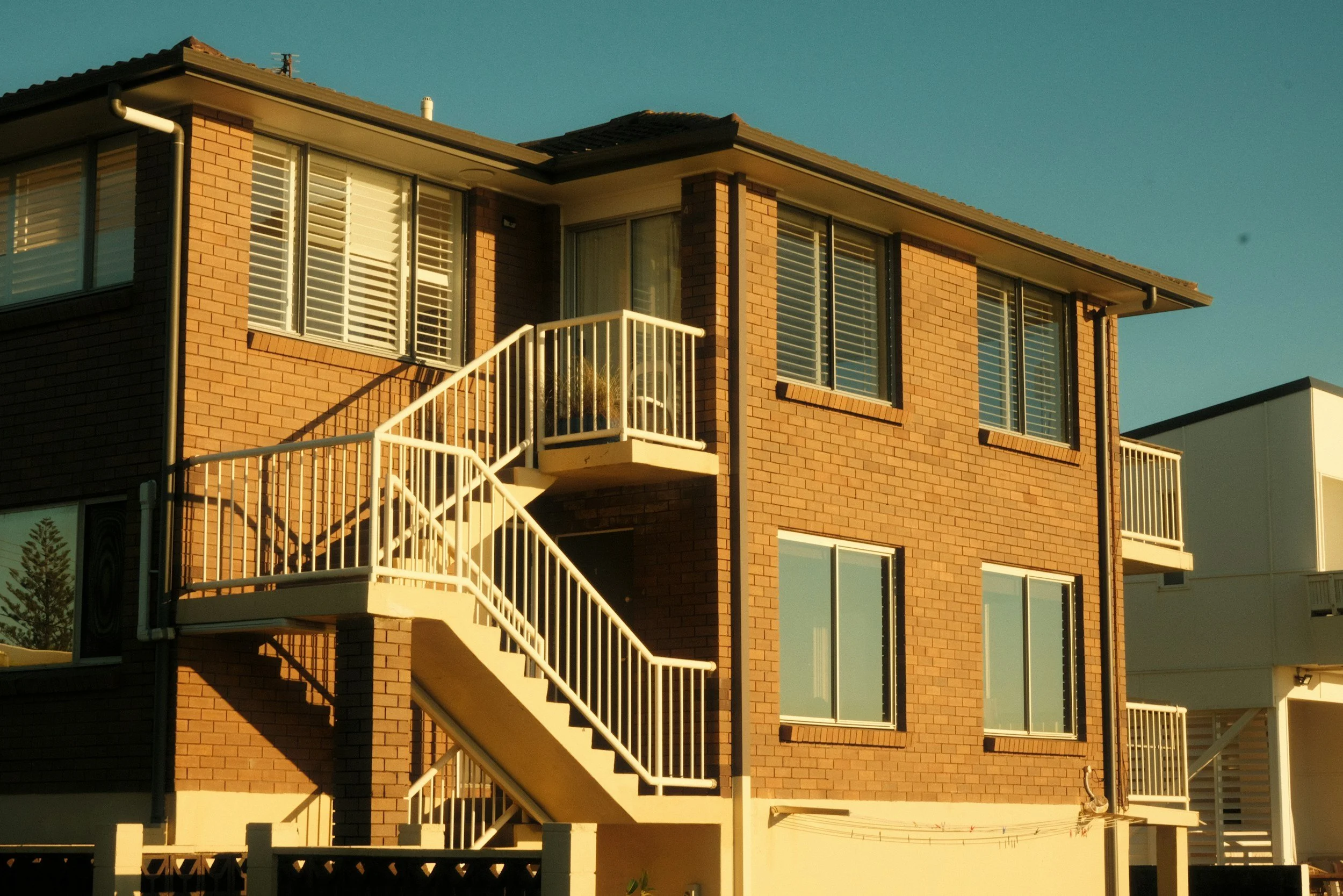 A multi-story brick residential building with white balcony staircases, large windows with white shutters, and a sloped roof, bathed in sunlight with a clear blue sky background.