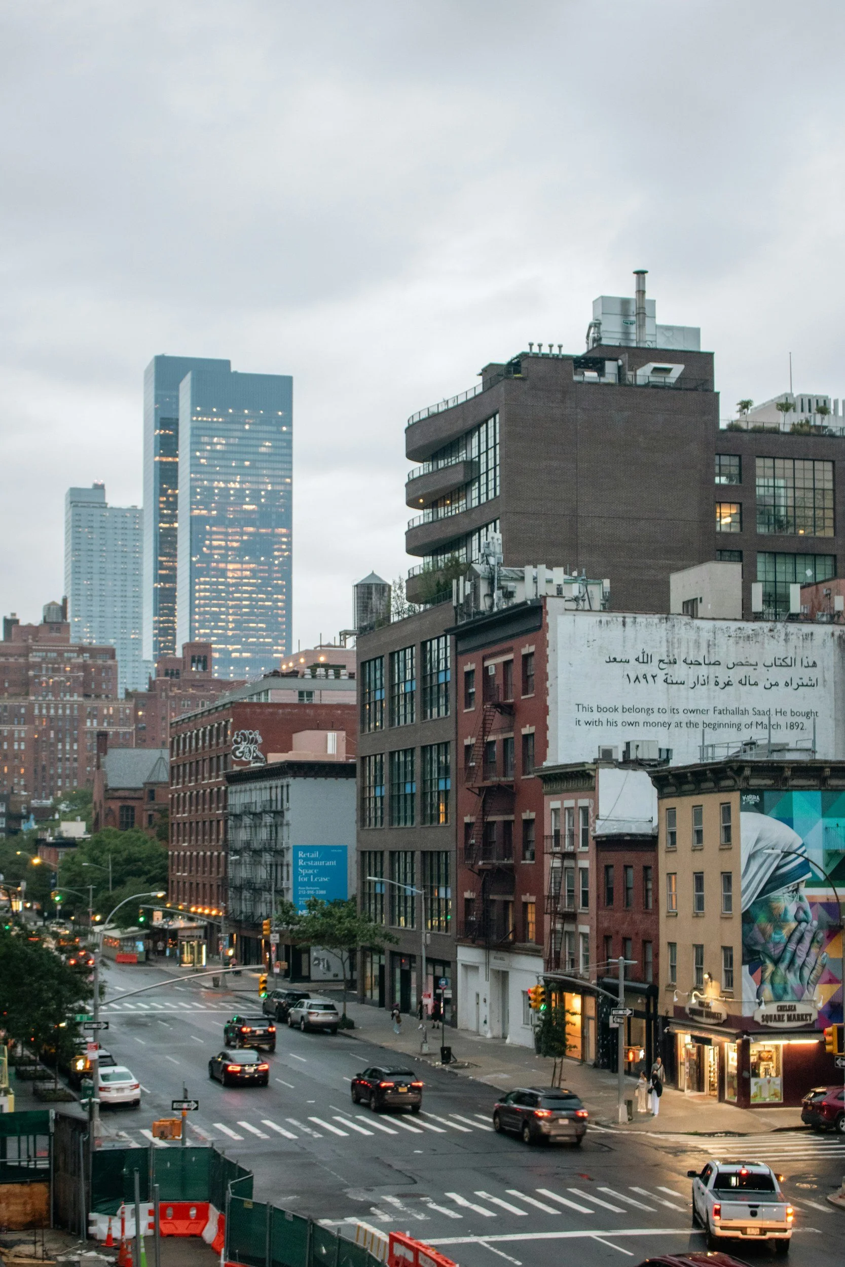 City street scene with cars and buildings, including a tall modern skyscraper, under a cloudy sky at dusk.