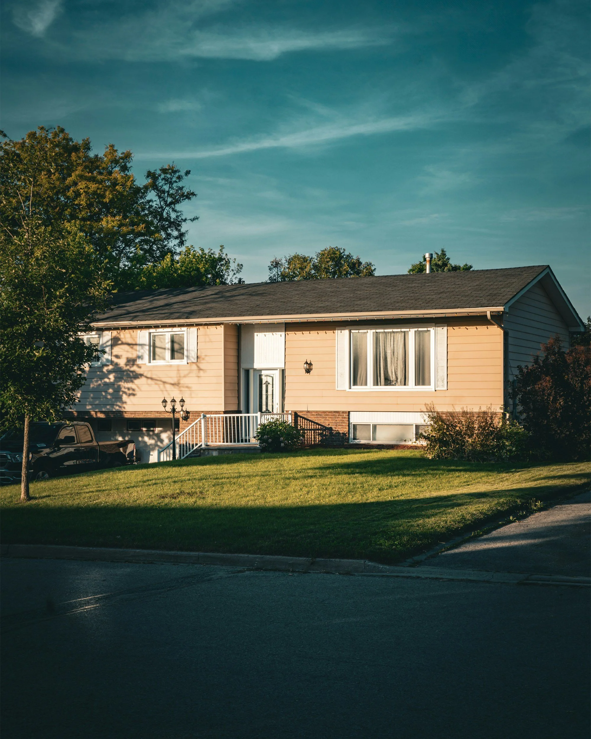 Residential house with fenced porch, parked car, lush green lawn, and trees under a blue sky with light clouds.
