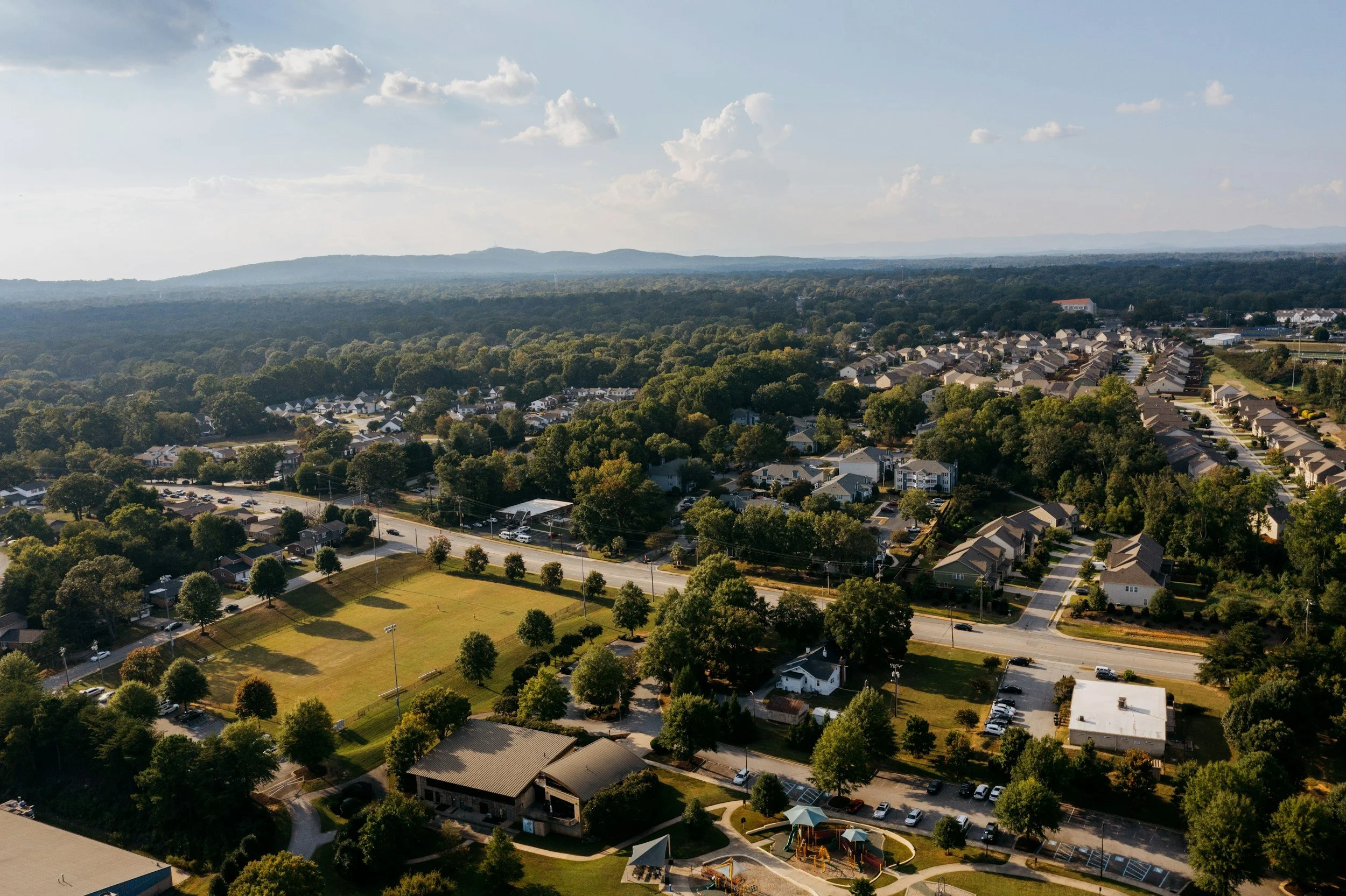 Aerial view of a suburban neighborhood with houses, trees, greenery, a baseball field, and a distant mountain range under a partly cloudy sky.