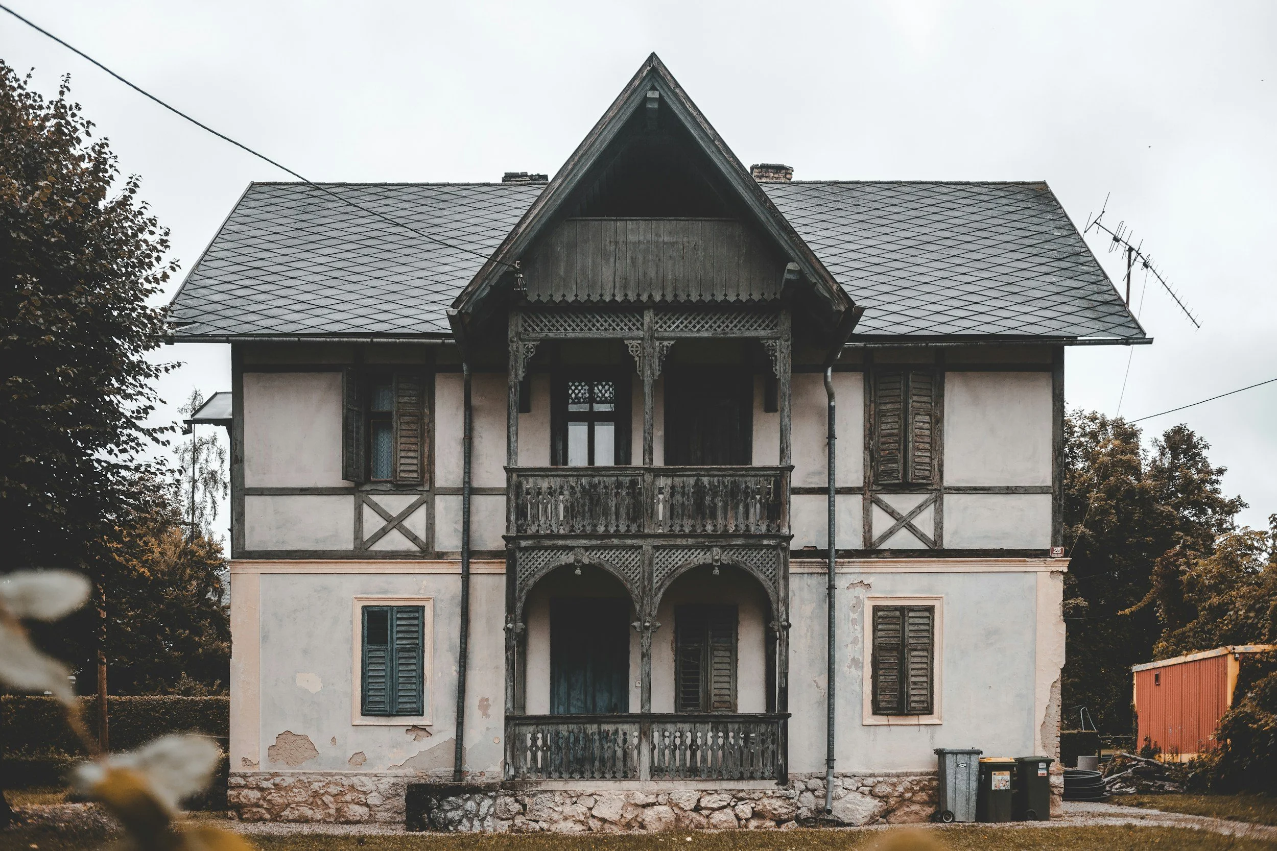 Old house with wooden balcony, shuttered windows, and weathered exterior, surrounded by trees.