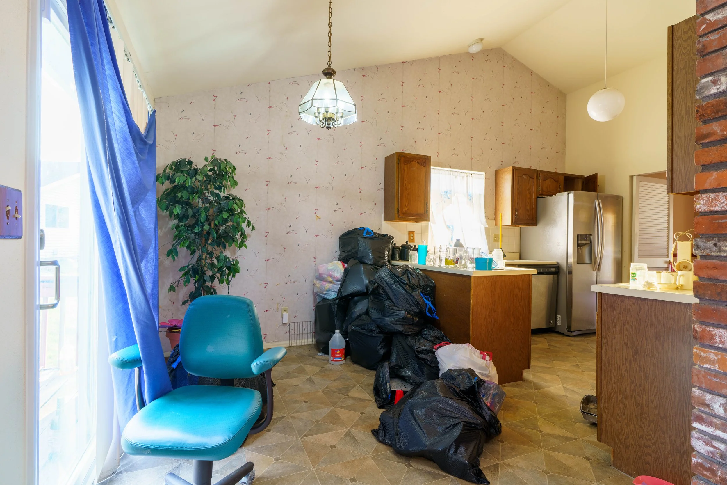 A cluttered kitchen with black trash bags on the floor, a teal chair, a window with white curtains, wooden kitchen cabinets, a refrigerator, and various items on the counter.