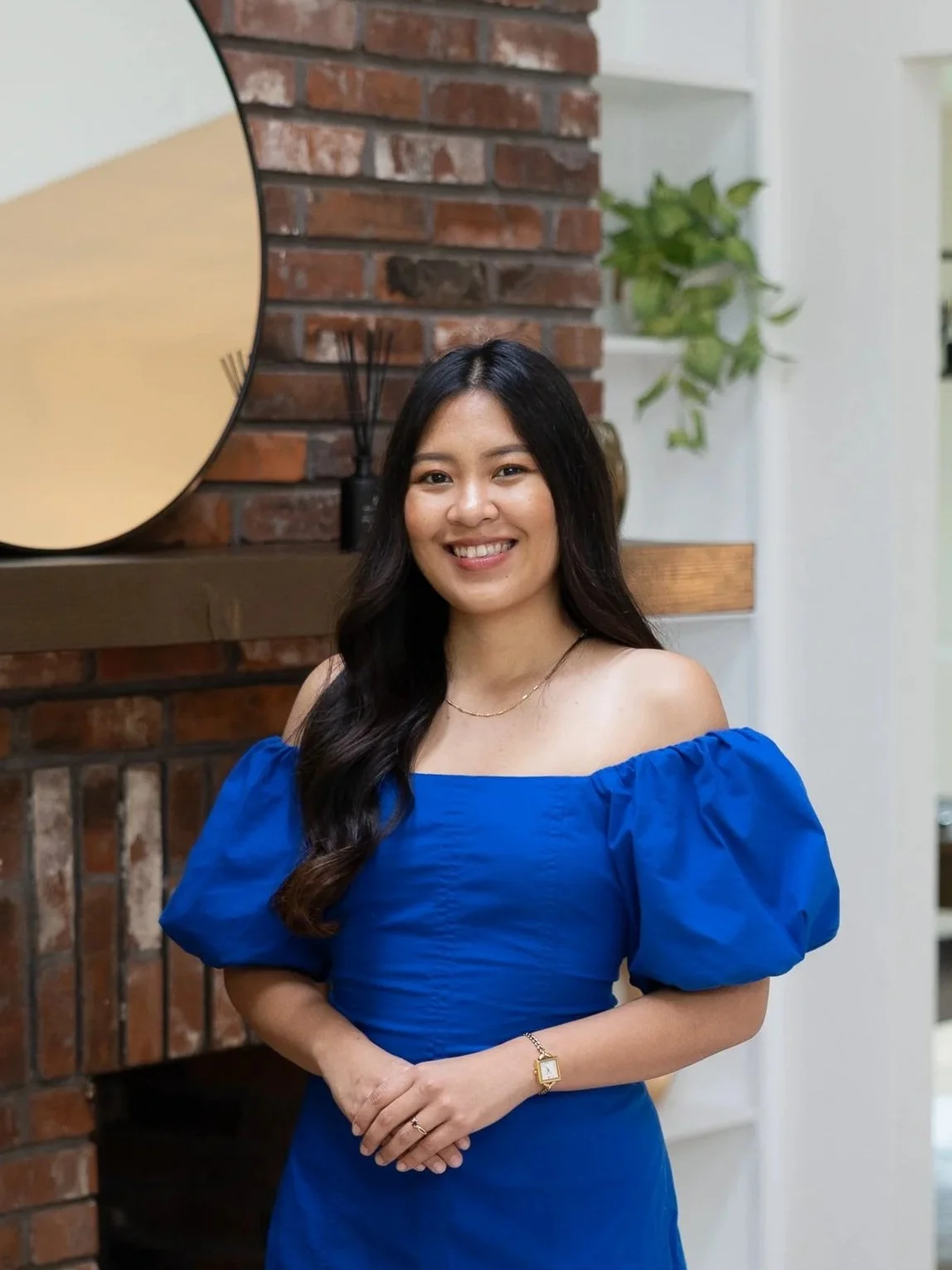 A woman with long dark hair wearing a bright blue off-shoulder dress, smiling, standing in front of a brick fireplace with a mirror and green plant in the background.