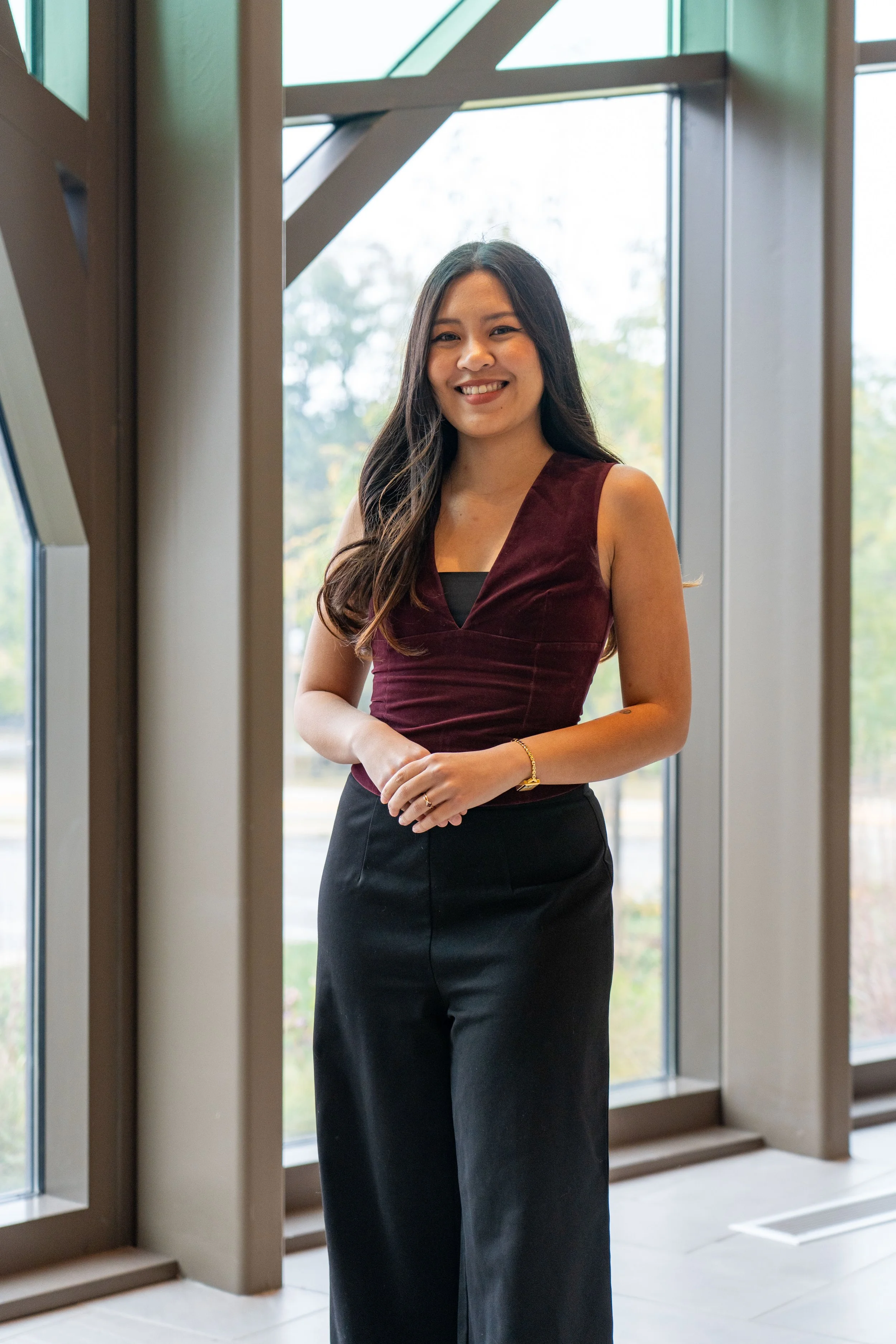 A young woman with long dark hair, wearing a sleeveless burgundy top and black wide-leg pants, standing inside near large windows with a view of trees outside, smiling at the camera.