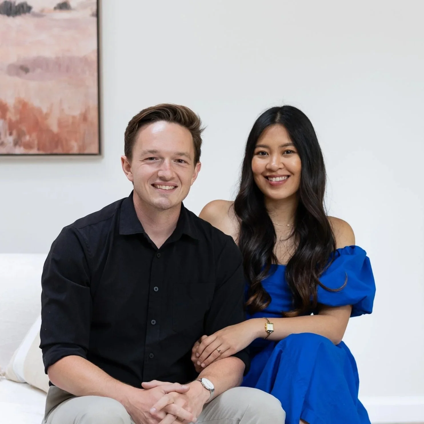 A man with brown hair and a woman with long dark hair sitting together in a room, both smiling. The man wears a black shirt and tan pants, and the woman wears a blue dress.