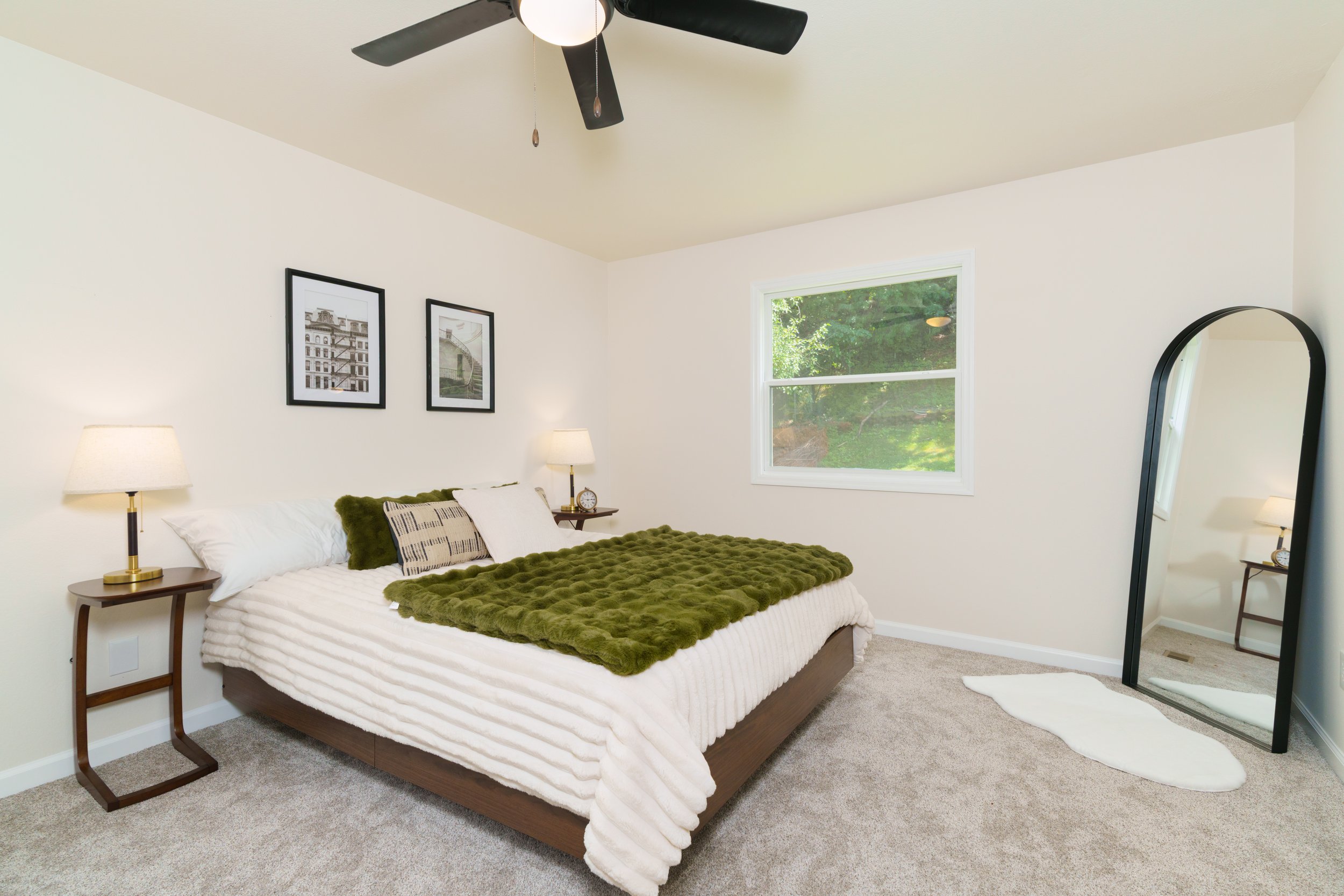 A bedroom with a bed covered in white and green bedding, two bedside lamps, framed black-and-white photographs on the wall, a window showing greenery outside, a full-length mirror, and beige carpeted floors.