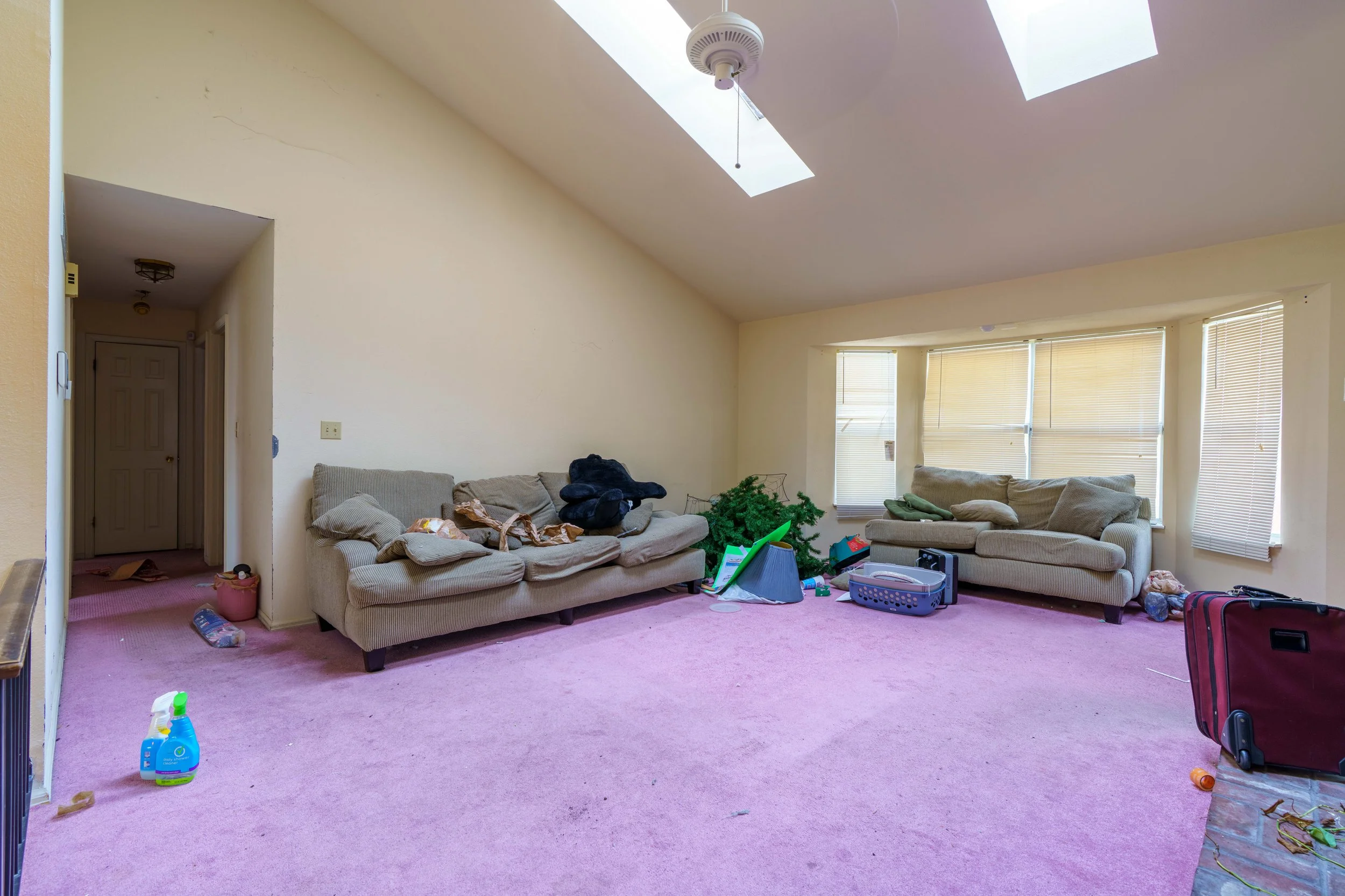 Living room with beige couches, cluttered with toys, clothes, and household items. Pink carpet, windows with beige blinds, ceiling fan, skylights, and a door visible.