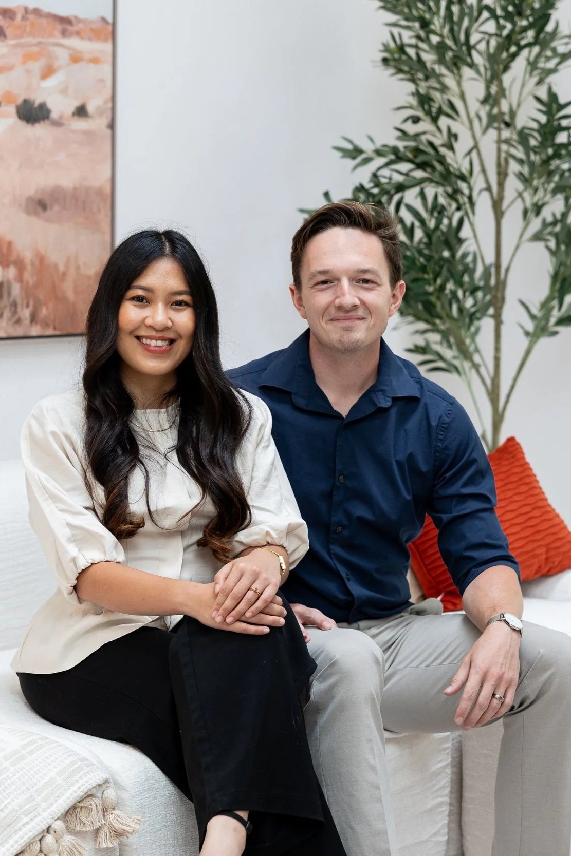 A woman with long dark hair and a man with short brown hair sitting on a white couch in a bright room with a green plant and abstract wall art behind them.