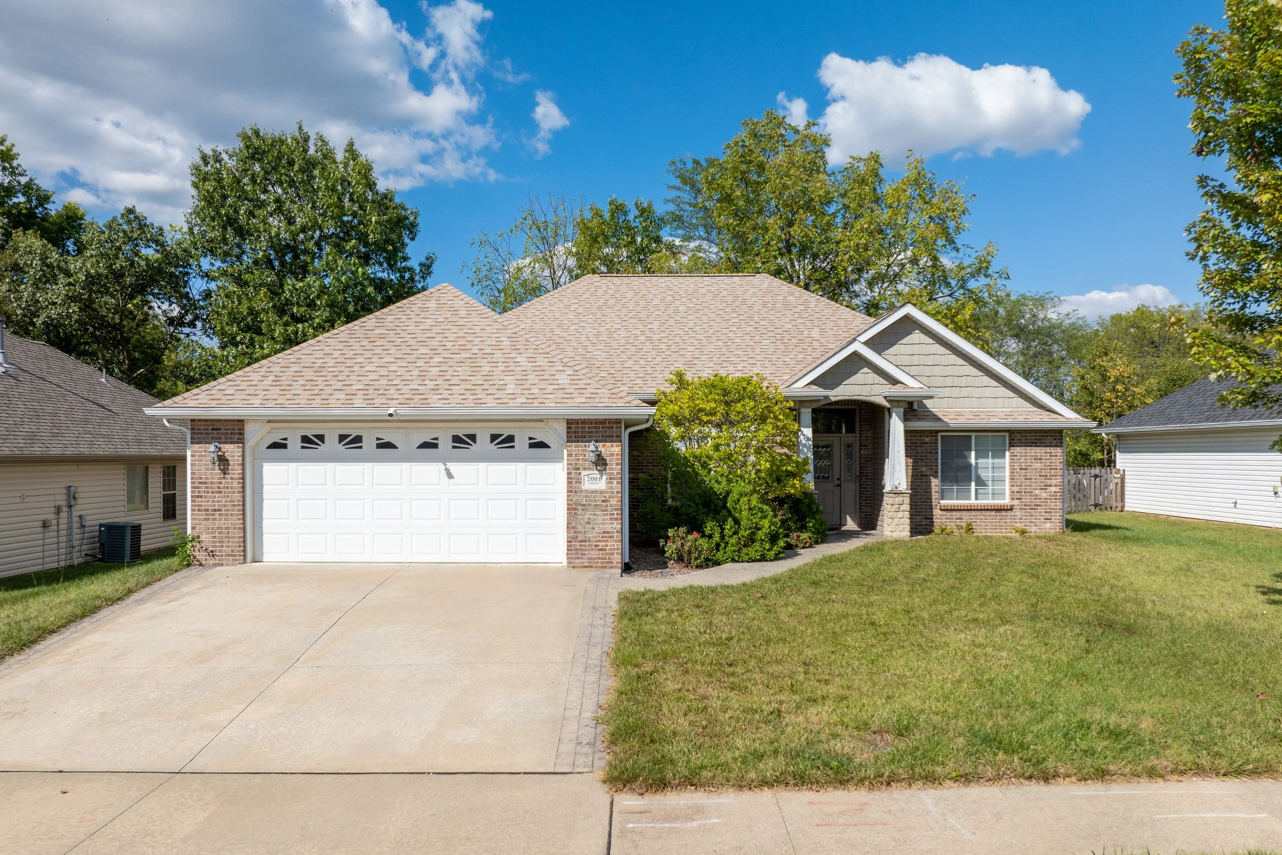 Front view of a suburban house with a brick facade, garage door, and a small front porch. The house has a tan-colored shingle roof, and there is well-maintained grass and a sidewalk in front. Trees and a blue sky with some clouds are visible in the background.