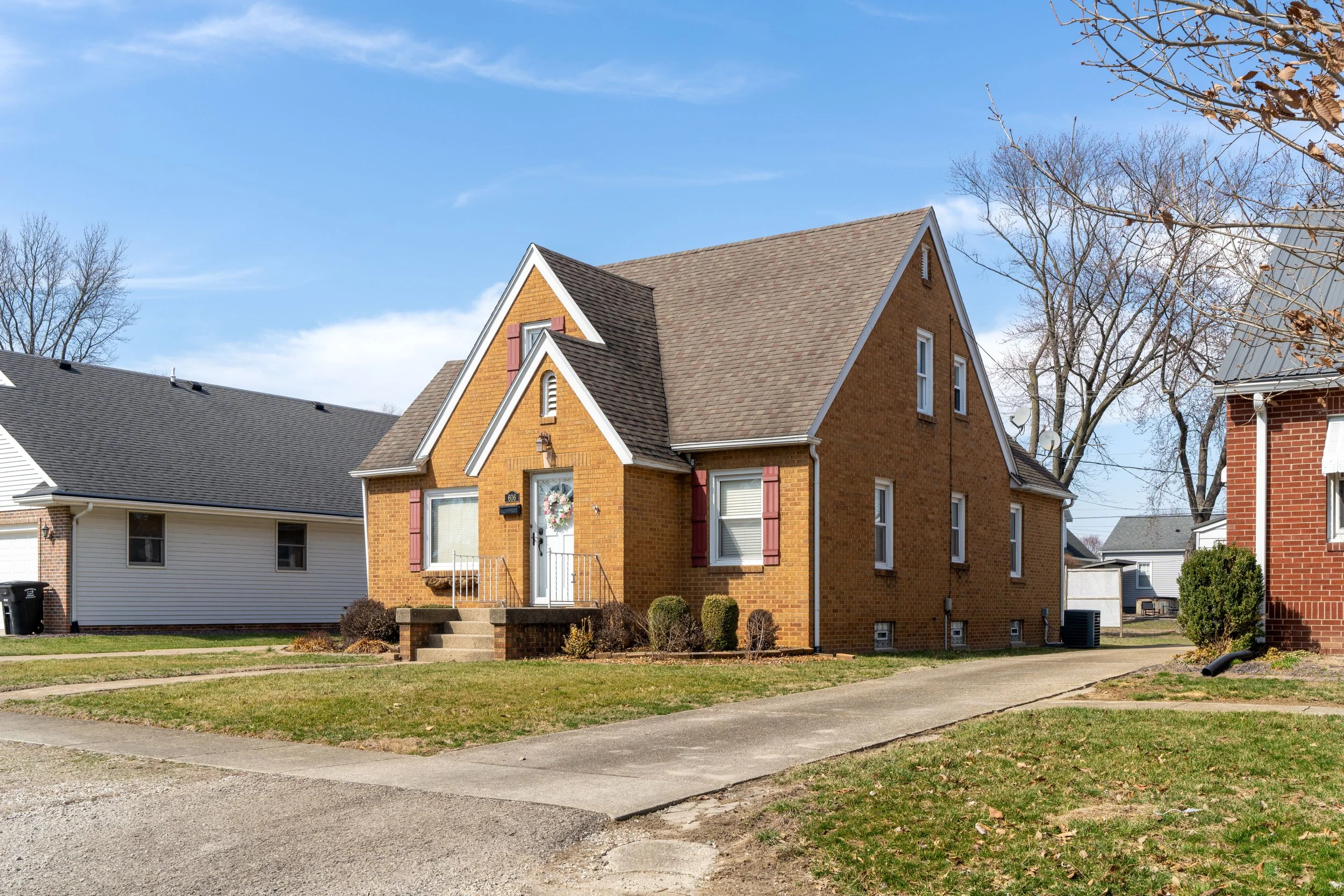 A two-story brick house with a tall, steeply-pitched gabled roof and white trim. The house has red shutters, a front door with a wreath, a small set of steps, and a front yard with grass and bushes. Surrounding houses and leafless trees are visible under a blue sky.