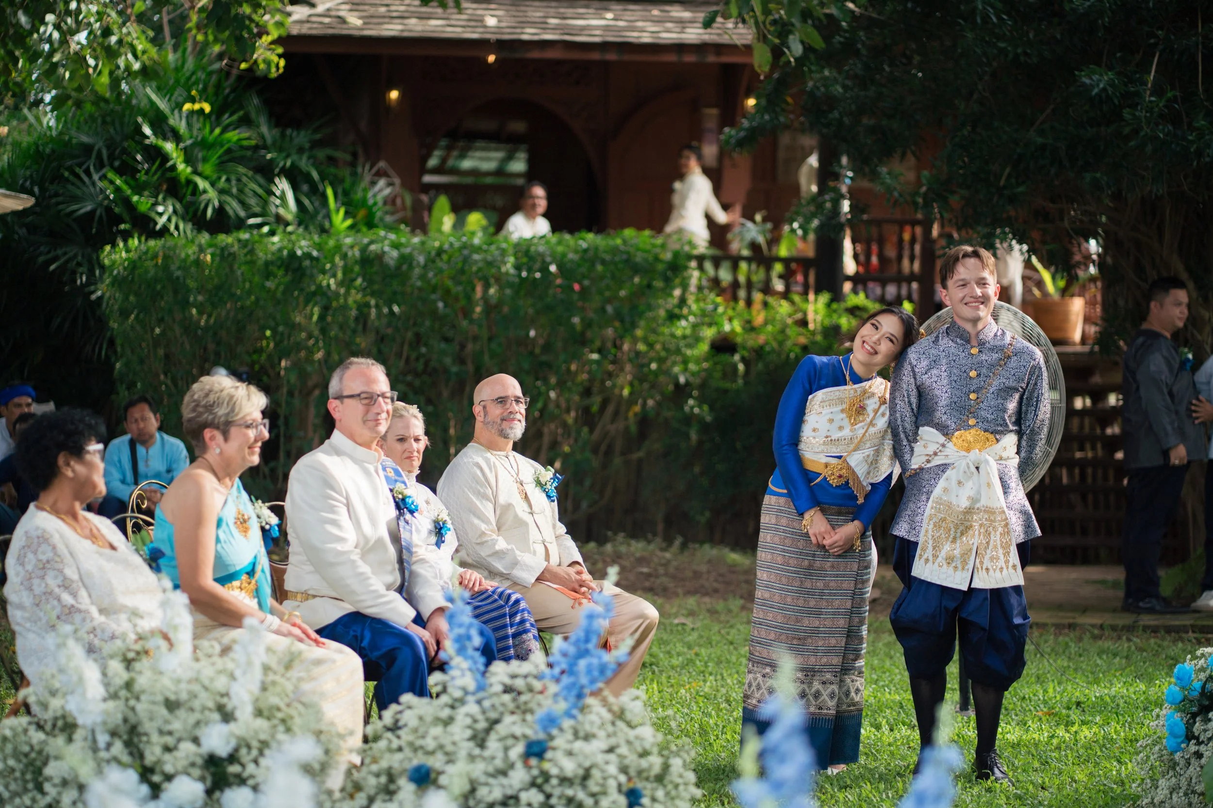 A diverse group of people at an outdoor wedding ceremony, with some seated and two standing in traditional attire, surrounded by greenery and flowers.