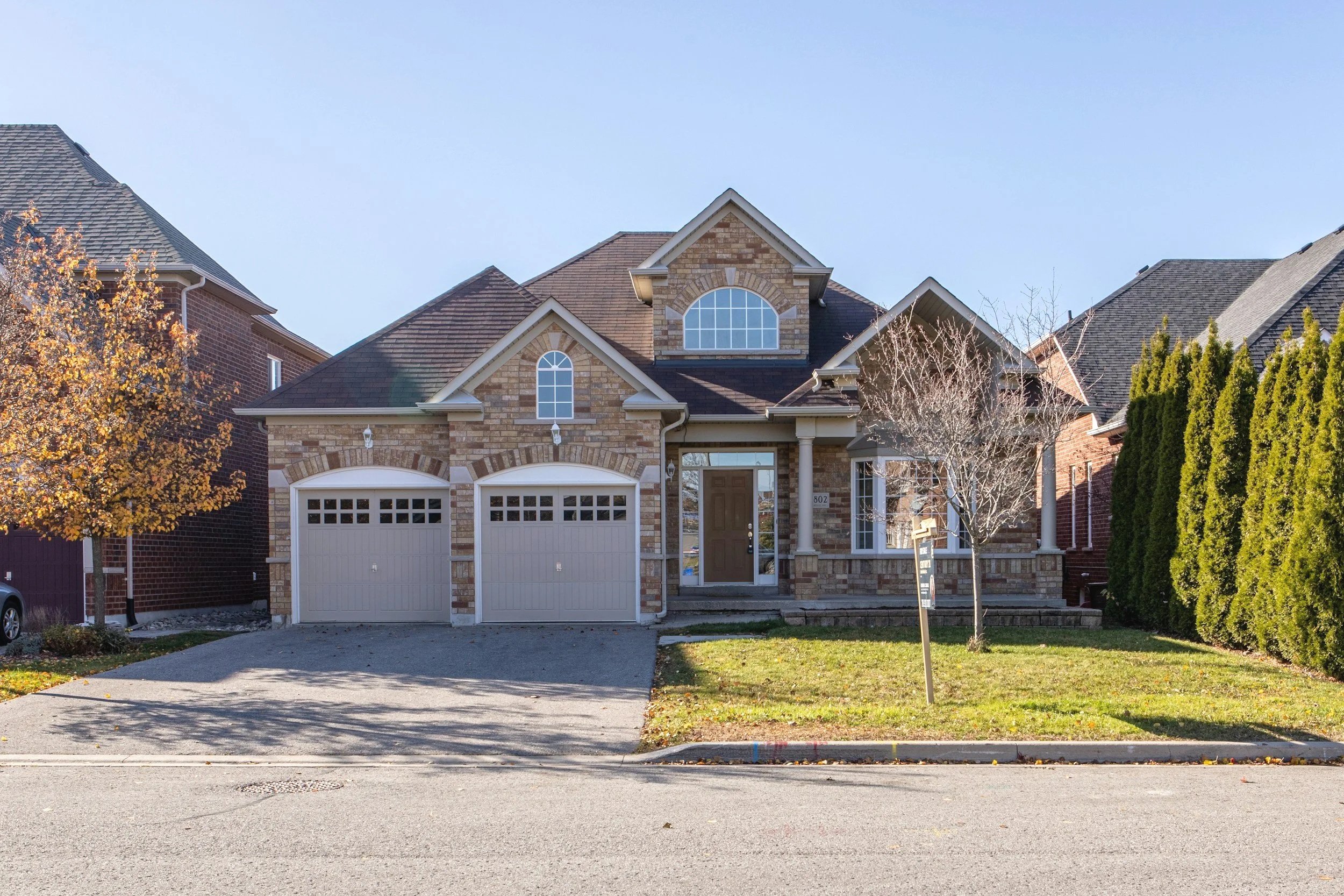 Front view of a two-story house with brick and stone exterior, white garage doors, brown front door, and a small front yard with a tree and lawn.