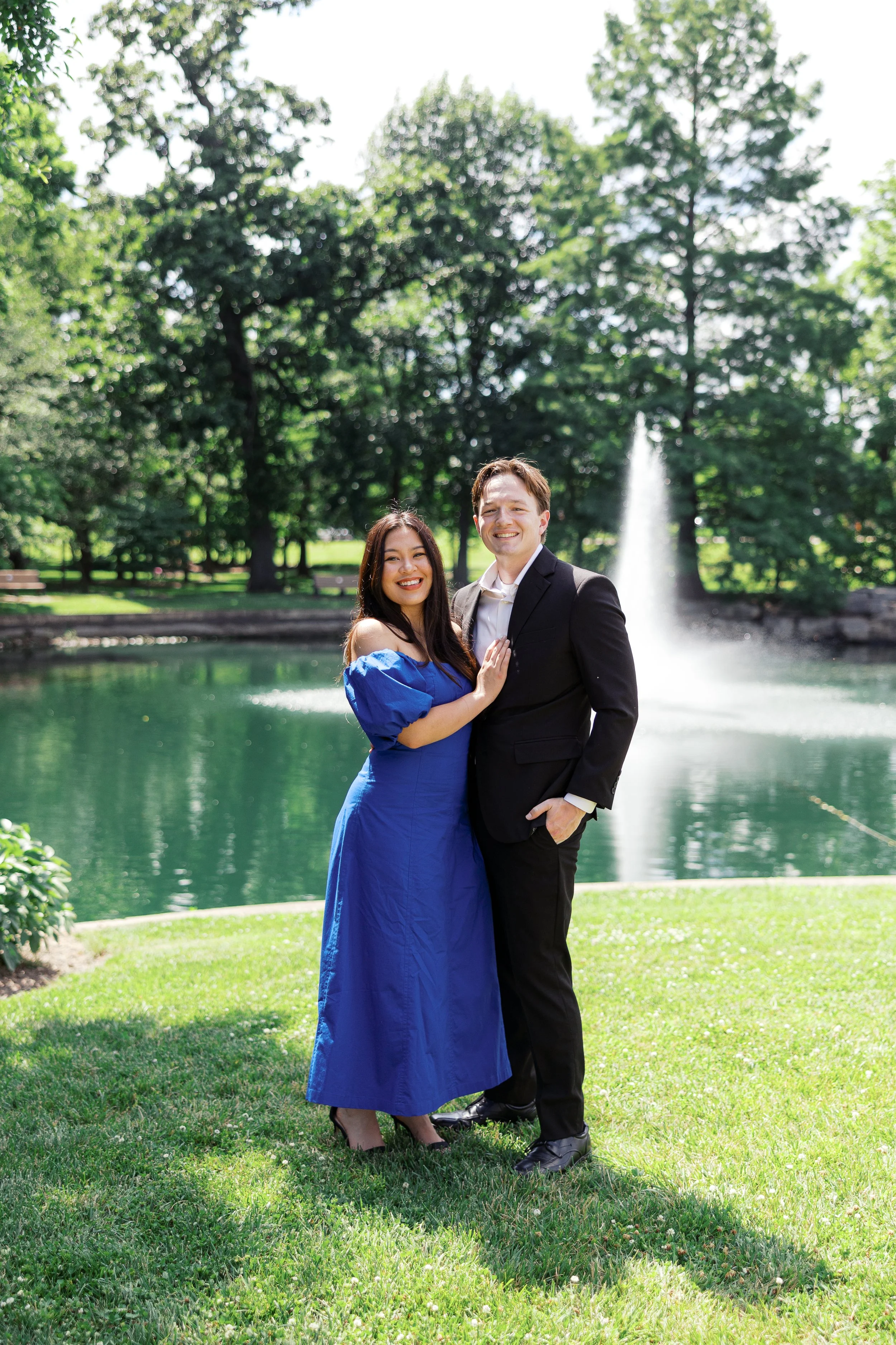 A young woman in a blue dress and a young man in a black suit are smiling and standing close together near a park pond with a fountain, green trees, and grass in the background.