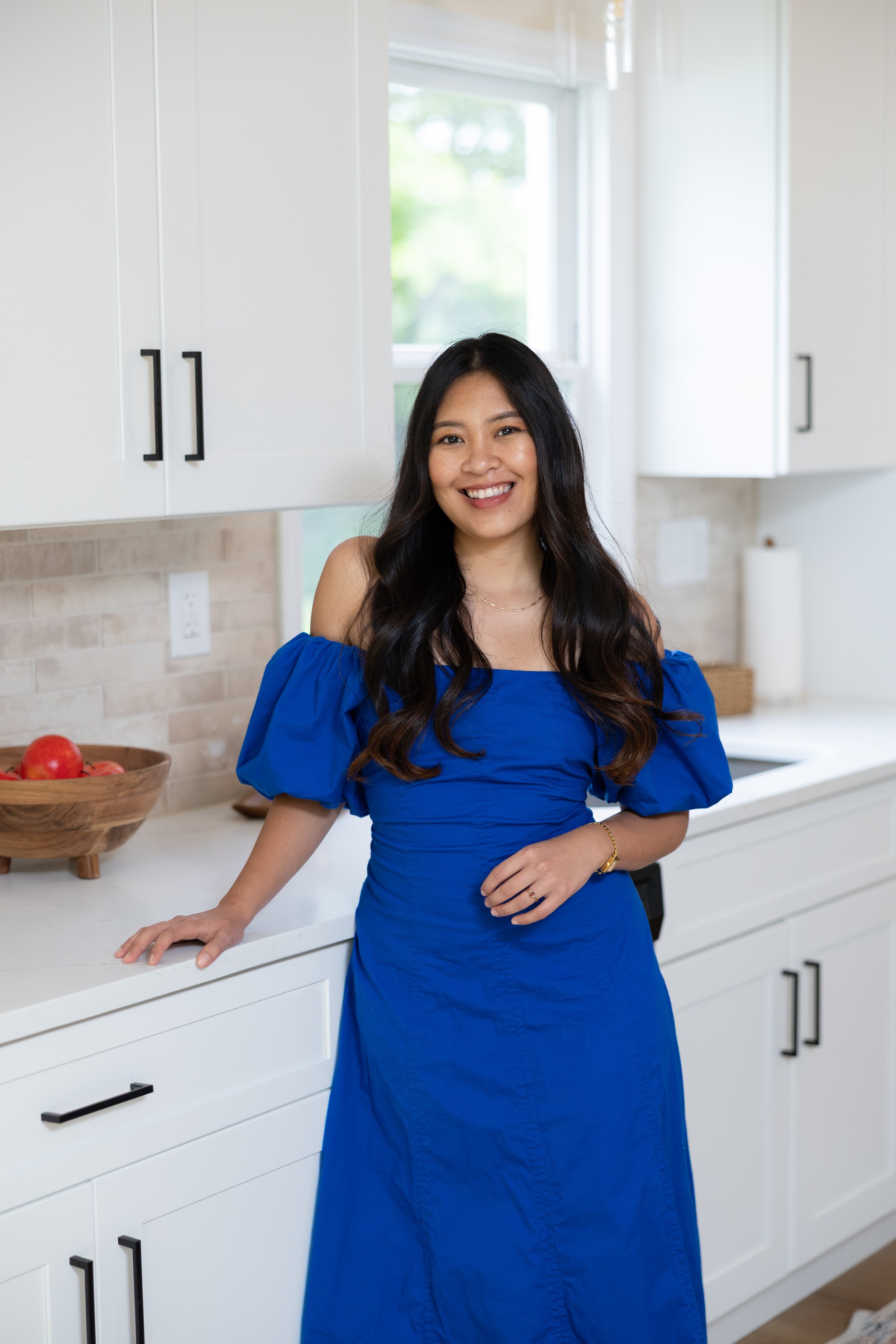 A woman in a royal blue dress standing in a modern kitchen with white cabinets and a wooden bowl of apples on the countertop, smiling at the camera.
