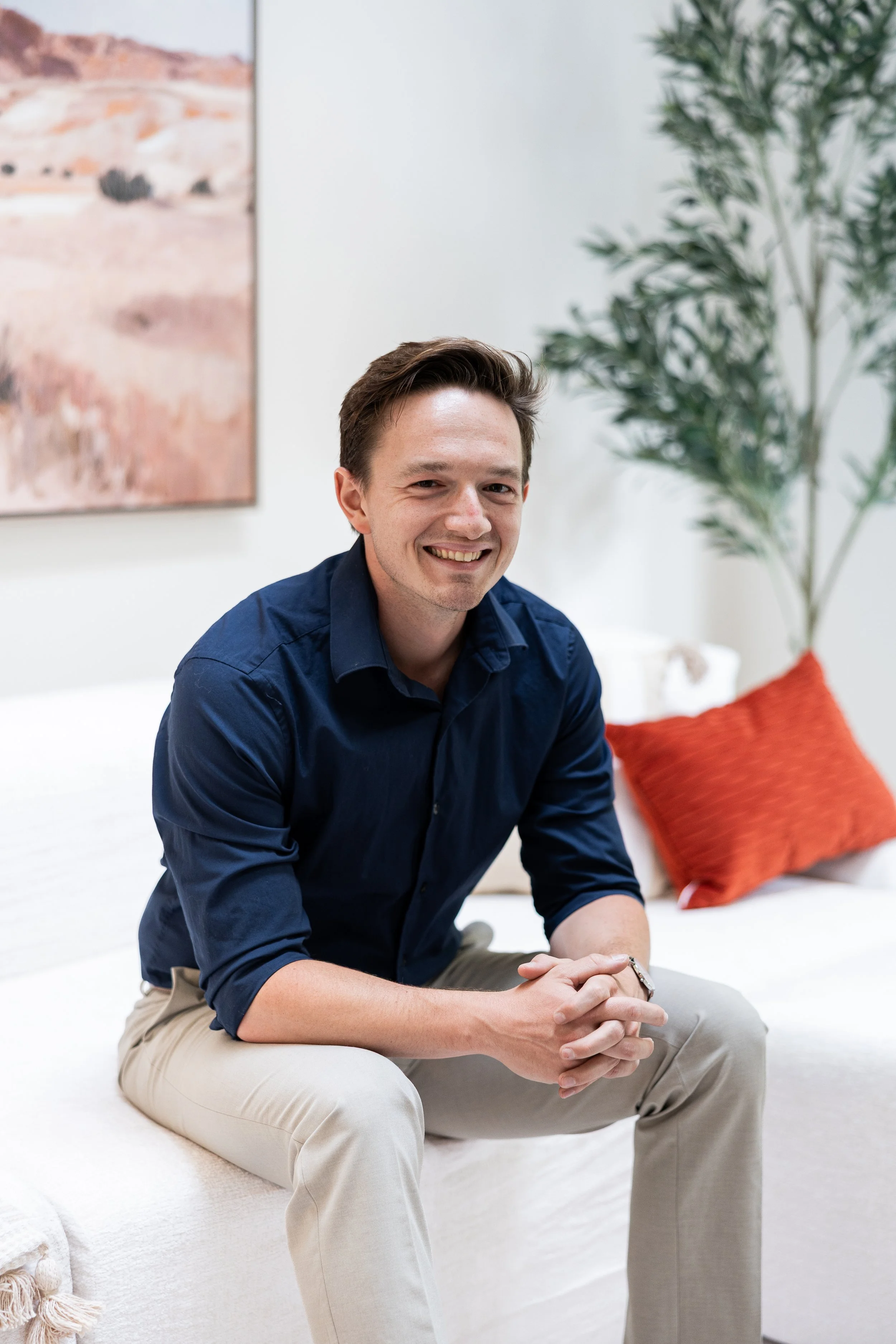A young man with short brown hair, wearing a navy blue button-up shirt and beige pants, is sitting on a white sofa, smiling at the camera. Behind him, there is a large green potted plant, a red pillow on the sofa, and a wall with an abstract painting.