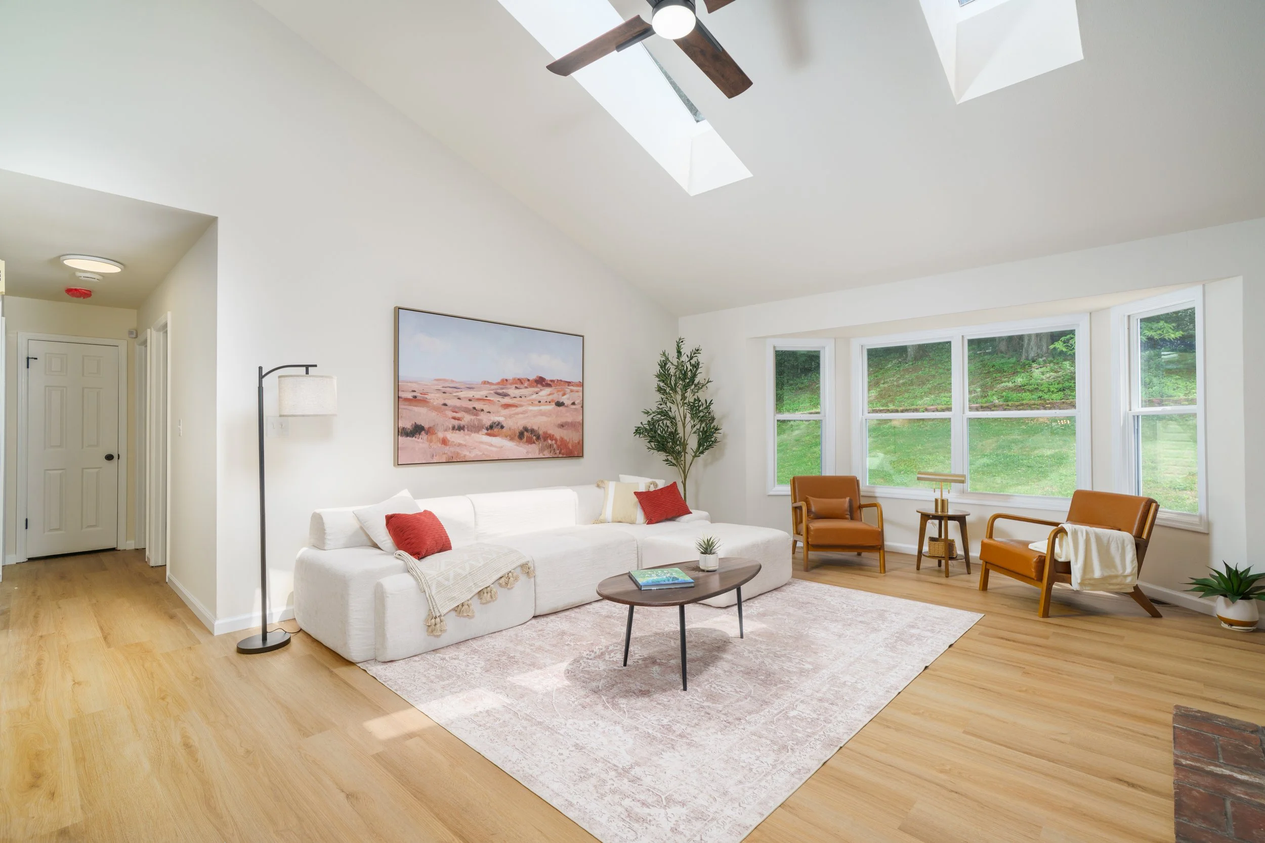 Living room with white sofas, red pillows, a side table, and several armchairs near large windows with a view of greenery outside. Skylights and a ceiling fan are visible on the ceiling.