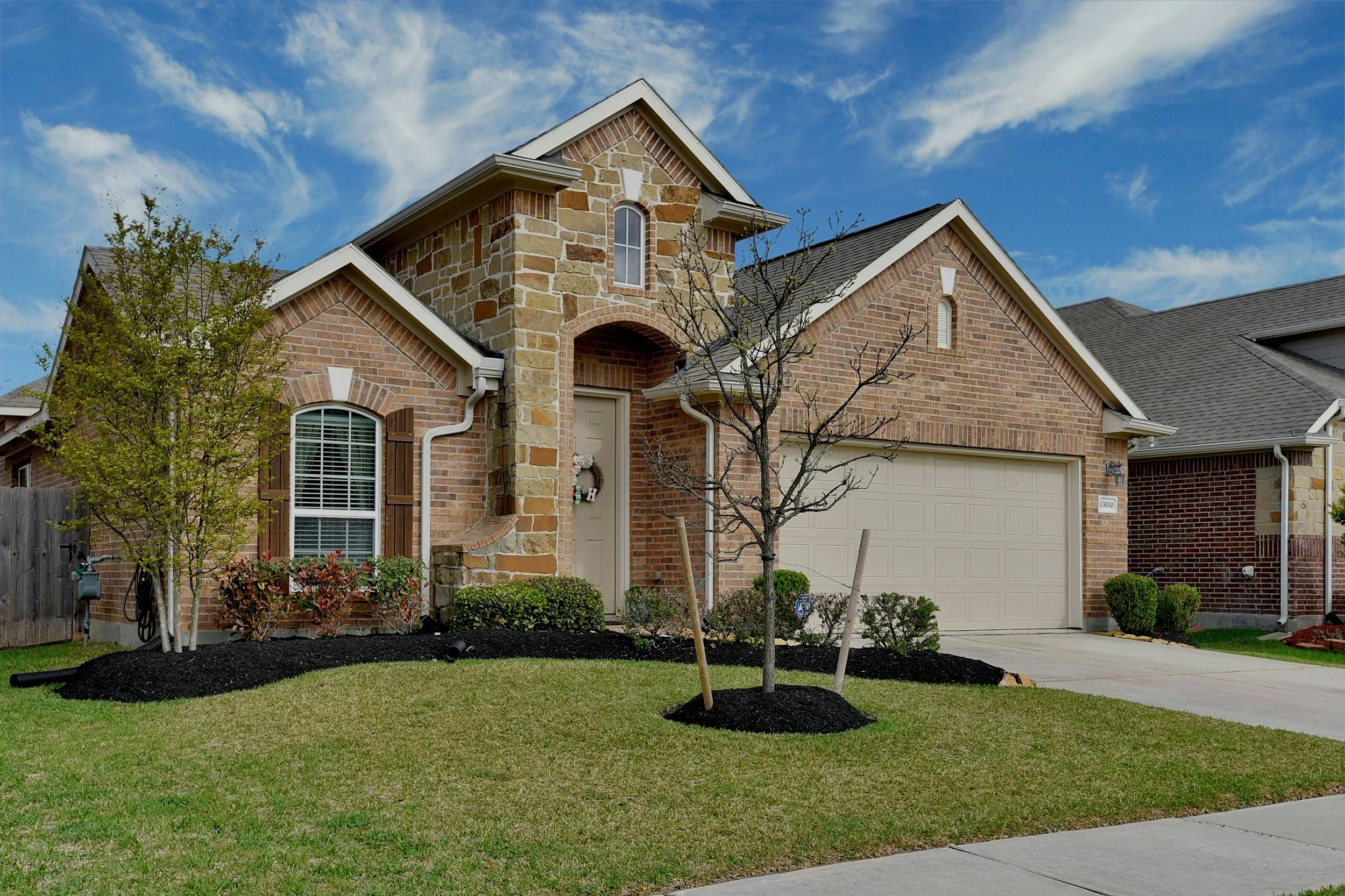 A two-story brick house with a garage, a front door decorated with a wreath, and leafless trees in the front yard. The house has a stone accent at the entryway, and there are small bushes and mulch in the yard. The sky is partly cloudy.