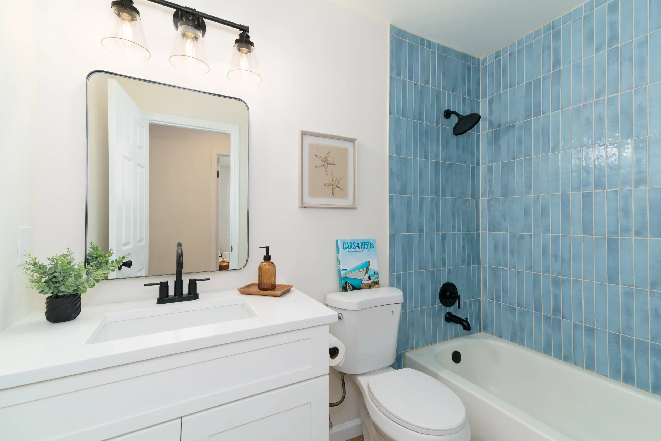 A modern bathroom featuring a white vanity with a black faucet, a large mirror, a potted plant, a soap dispenser, a framed picture of starfish, and a bathtub with blue tiled wall behind it. The showerhead is mounted on the tiled wall, and a book titled "CARS of the 1950s" is on the toilet tank.