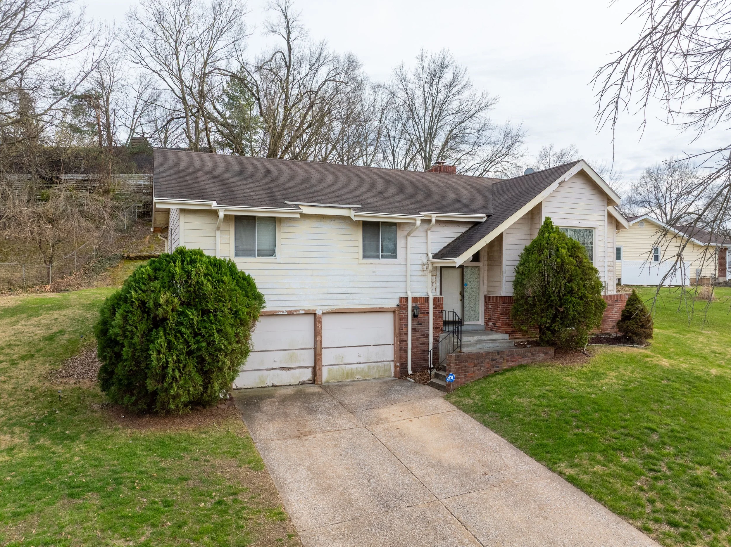 Front view of a two-story suburban house with a white and brick exterior, small front porch, green bushes, grass lawn, sidewalk, and leafless trees in the background under cloudy sky.