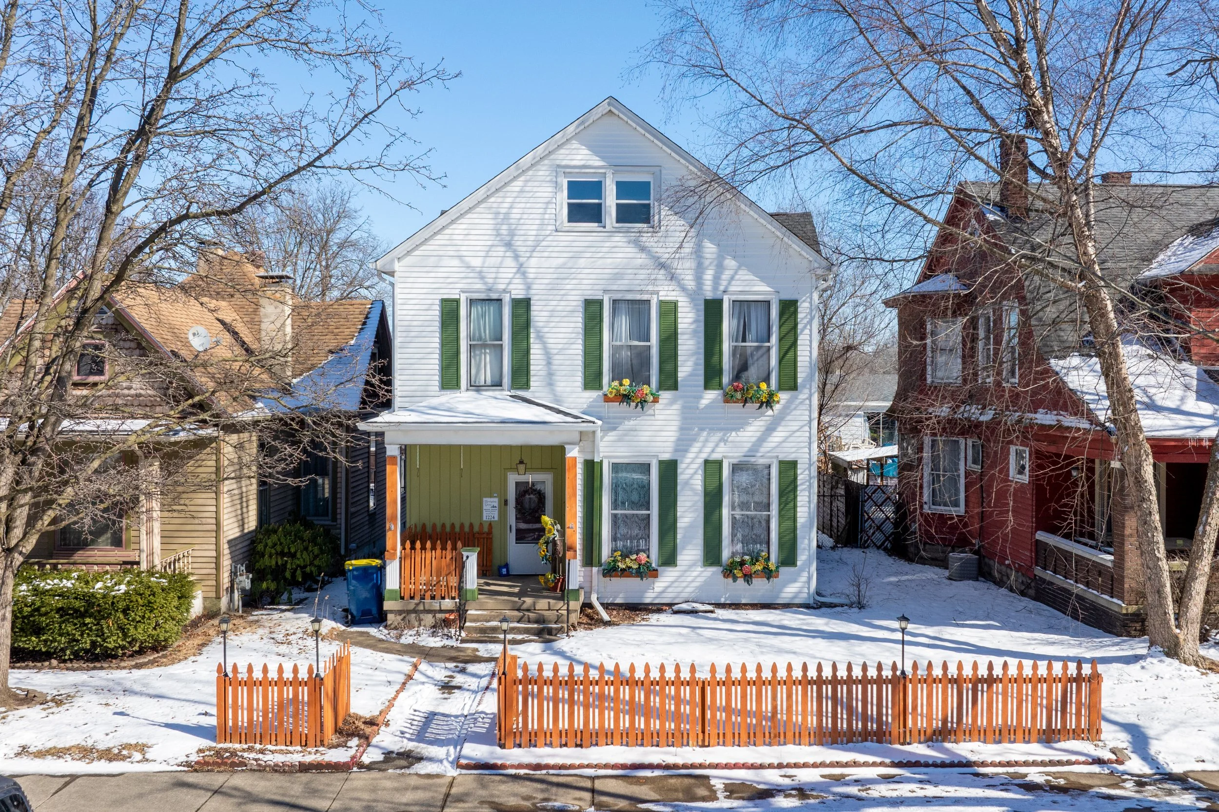A white house with green shutters and flower boxes, in a snowy neighborhood, with leafless trees and neighboring houses.