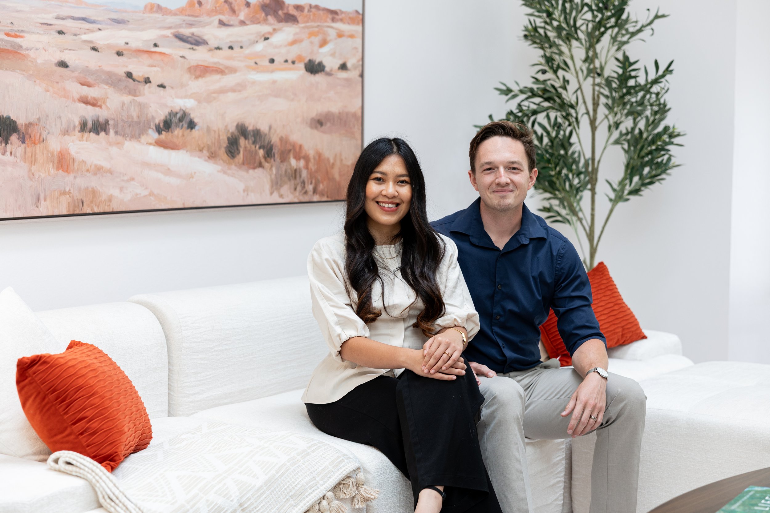 A man and a woman sitting together on a white couch in a modern room. The woman has long black hair, is smiling, and wears a light-colored top. The man has short brown hair, is smiling, and wears a dark blue shirt. There are orange pillows on the couch, a large abstract painting on the wall behind them, and a tall green plant in the corner.