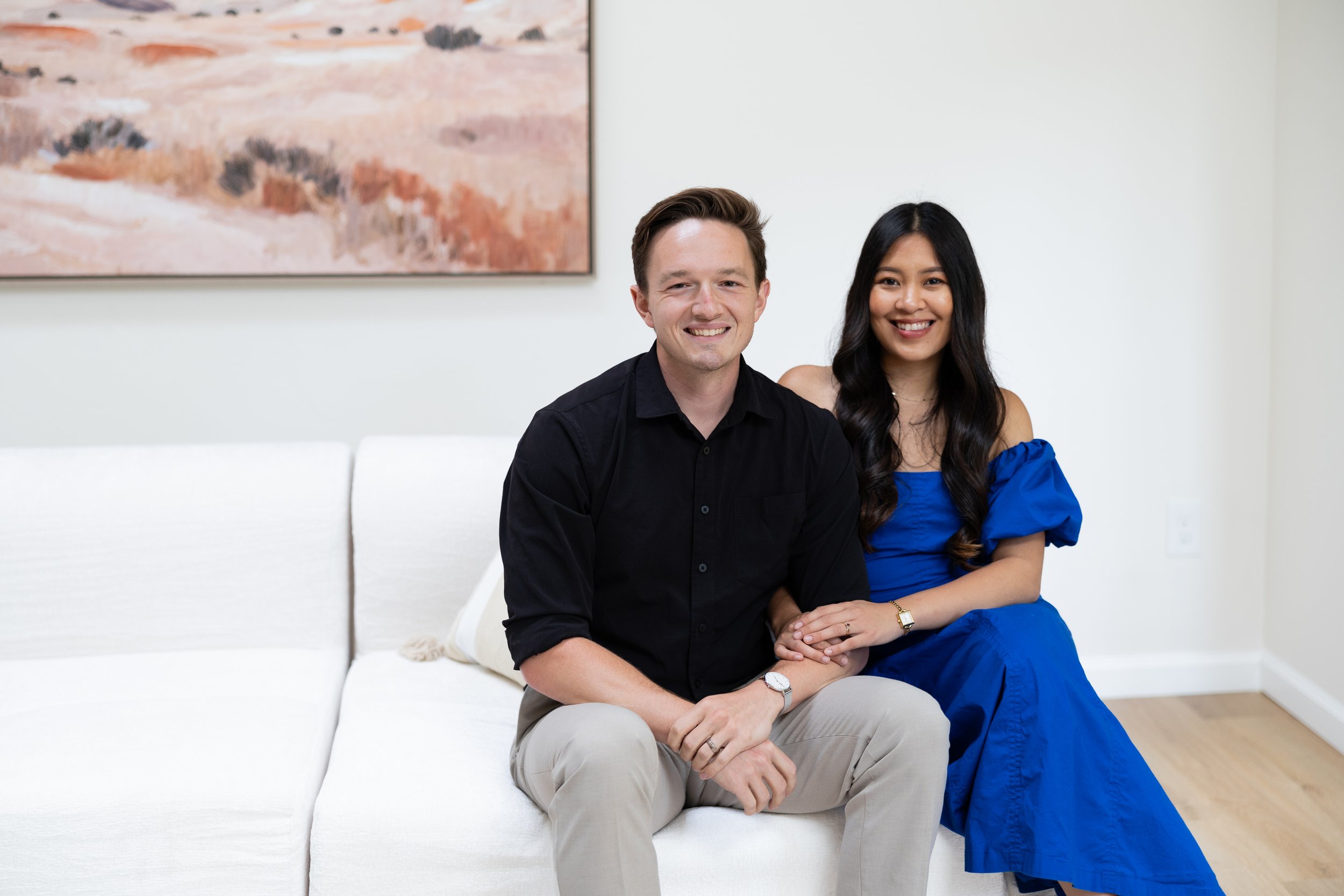 A smiling man with short brown hair wearing a black shirt and beige pants, sitting with a woman with long black hair wearing a blue dress, sitting on a white couch in a room with plain white walls and a framed abstract painting in the background.