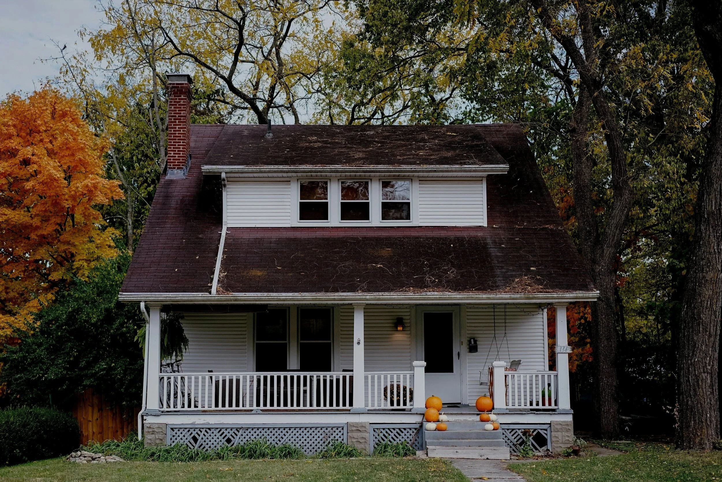 A two-story white house with a porch, surrounded by trees with fall foliage. The house has a steep roof with a chimney, and pumpkins are placed on the steps leading to the porch.