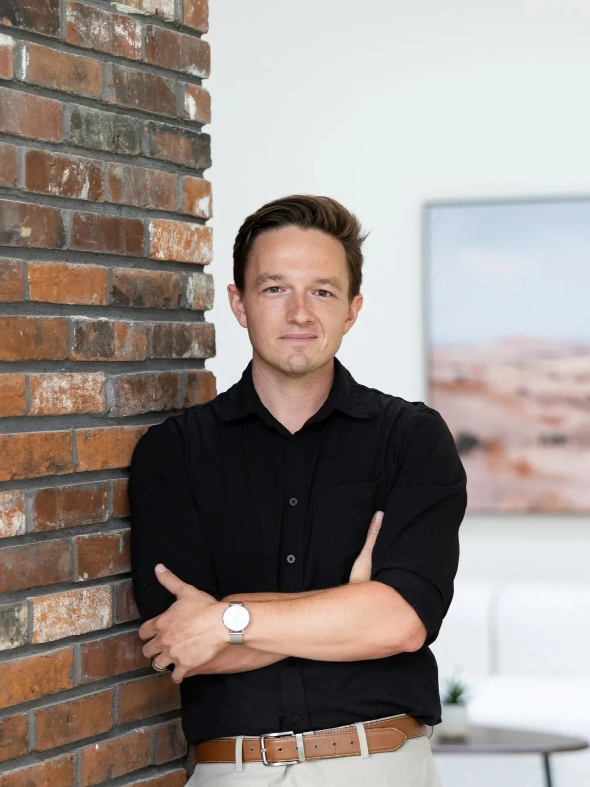 A man with light skin and brown hair stands with arms crossed, leaning against a brick wall in a modern indoor setting. He wears a black button-down shirt and a silver watch.