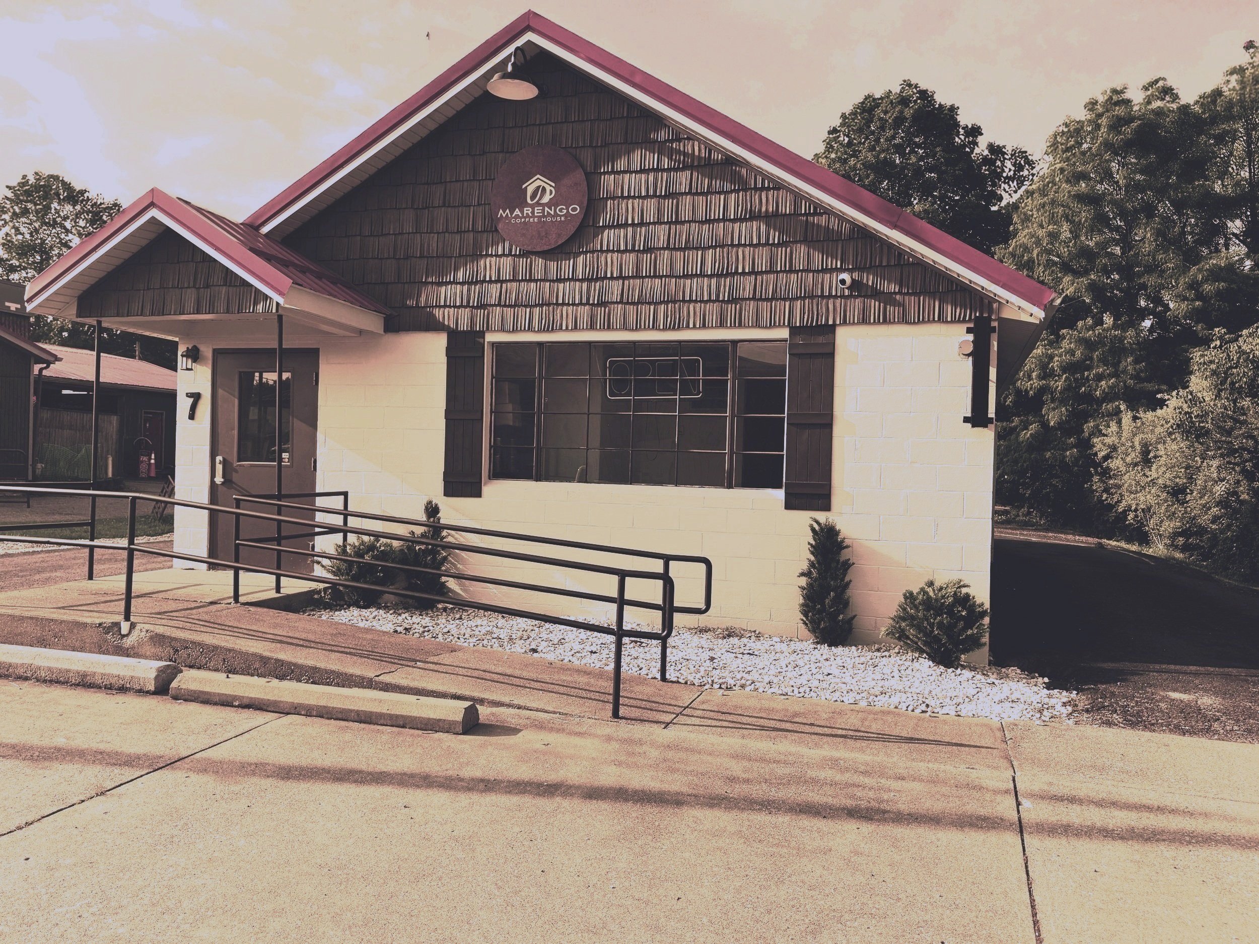 Coffee shop named Marengo Coffee House with a dark brown shingled upper exterior, cream-colored brick lower exterior, large front window, black shutters, outdoor ramp, and small landscaping with bushes and white gravel, surrounded by trees.