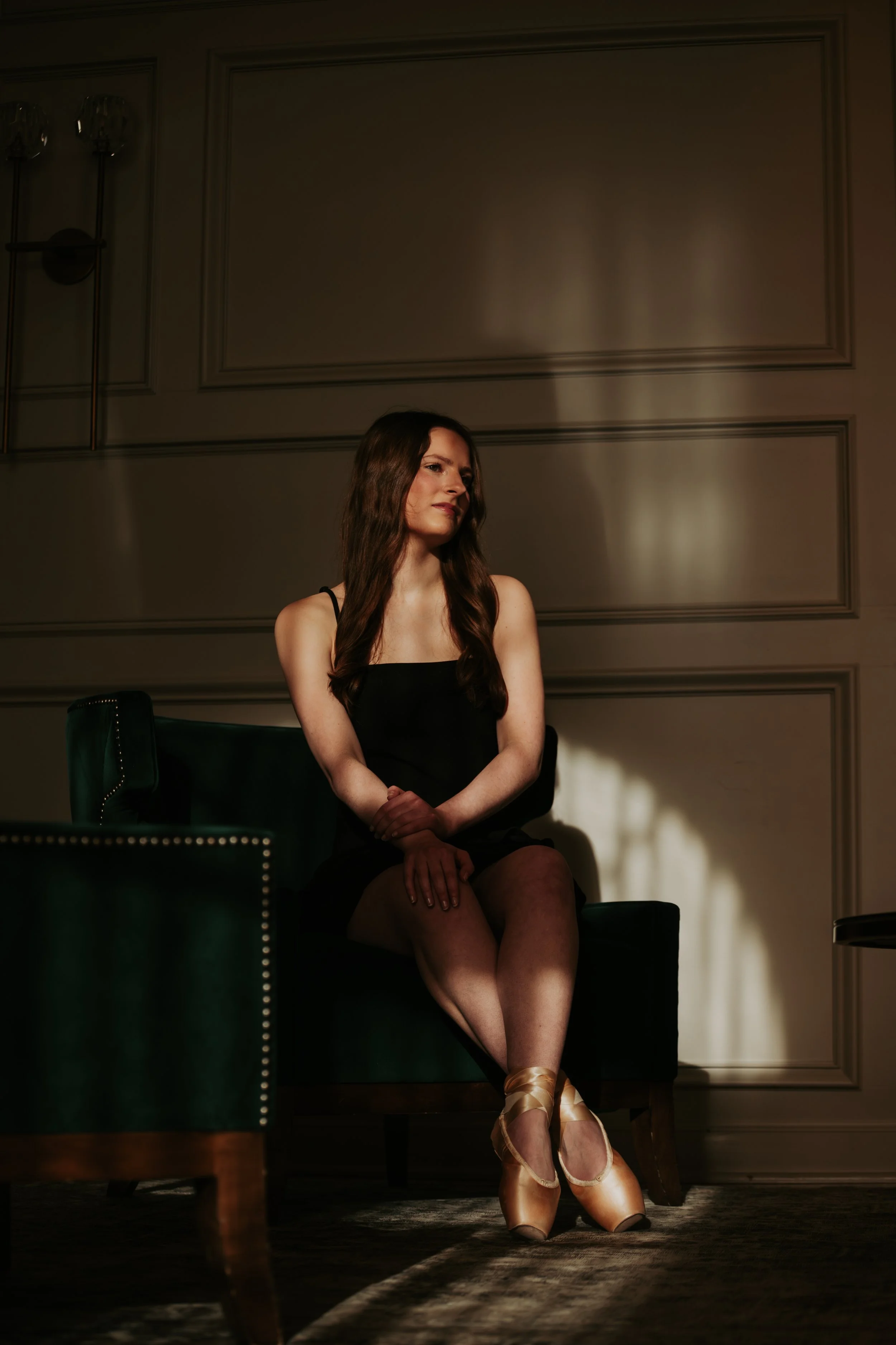 A woman in a black dress with long brown hair sitting on a dark green armchair in a dimly lit room with decorative wall paneling, illuminated by a focused light source.