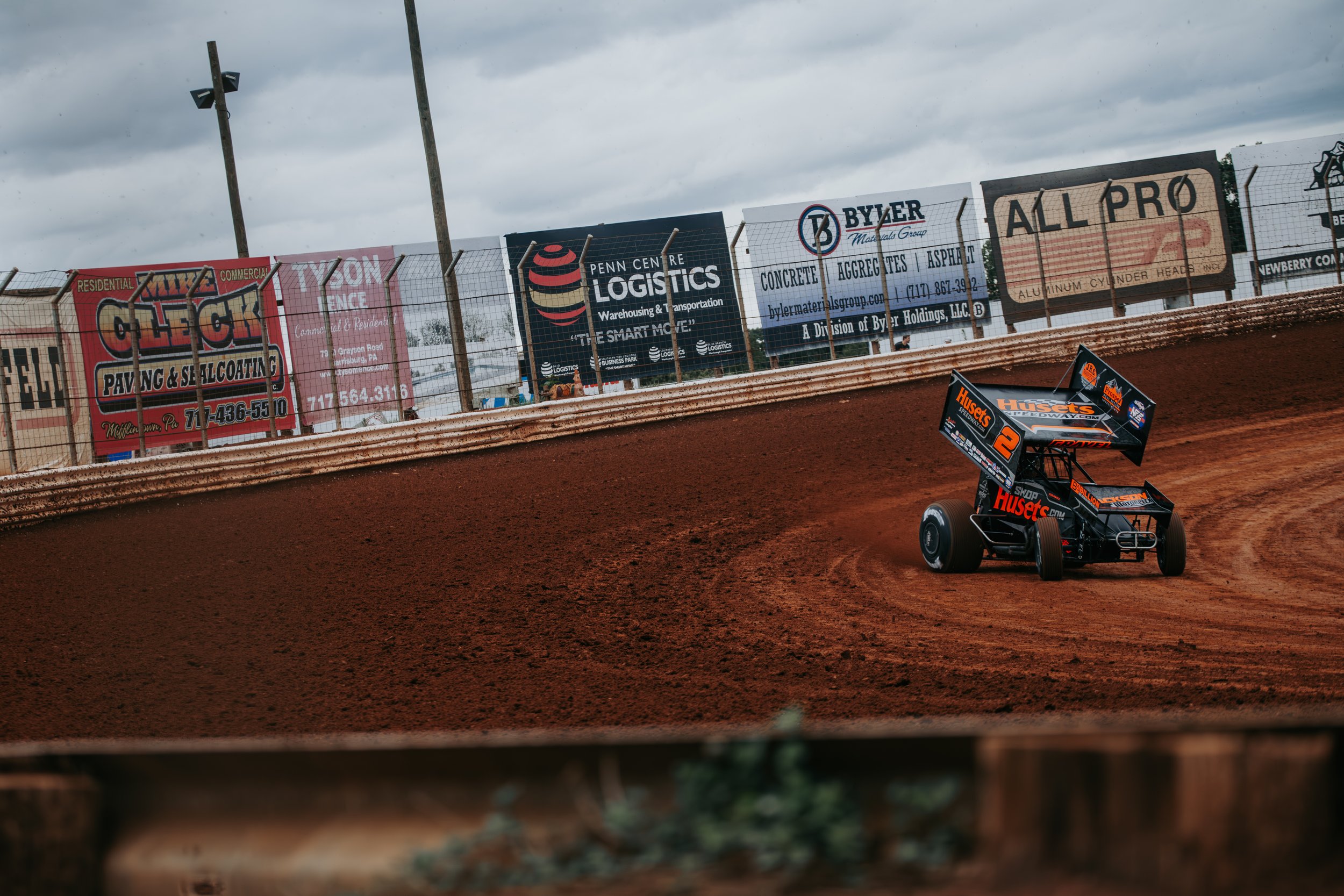 A dirt race track with a sprint car speeding around a corner, seen through a gap in a wooden fence. The background features green trees and a blue sky.