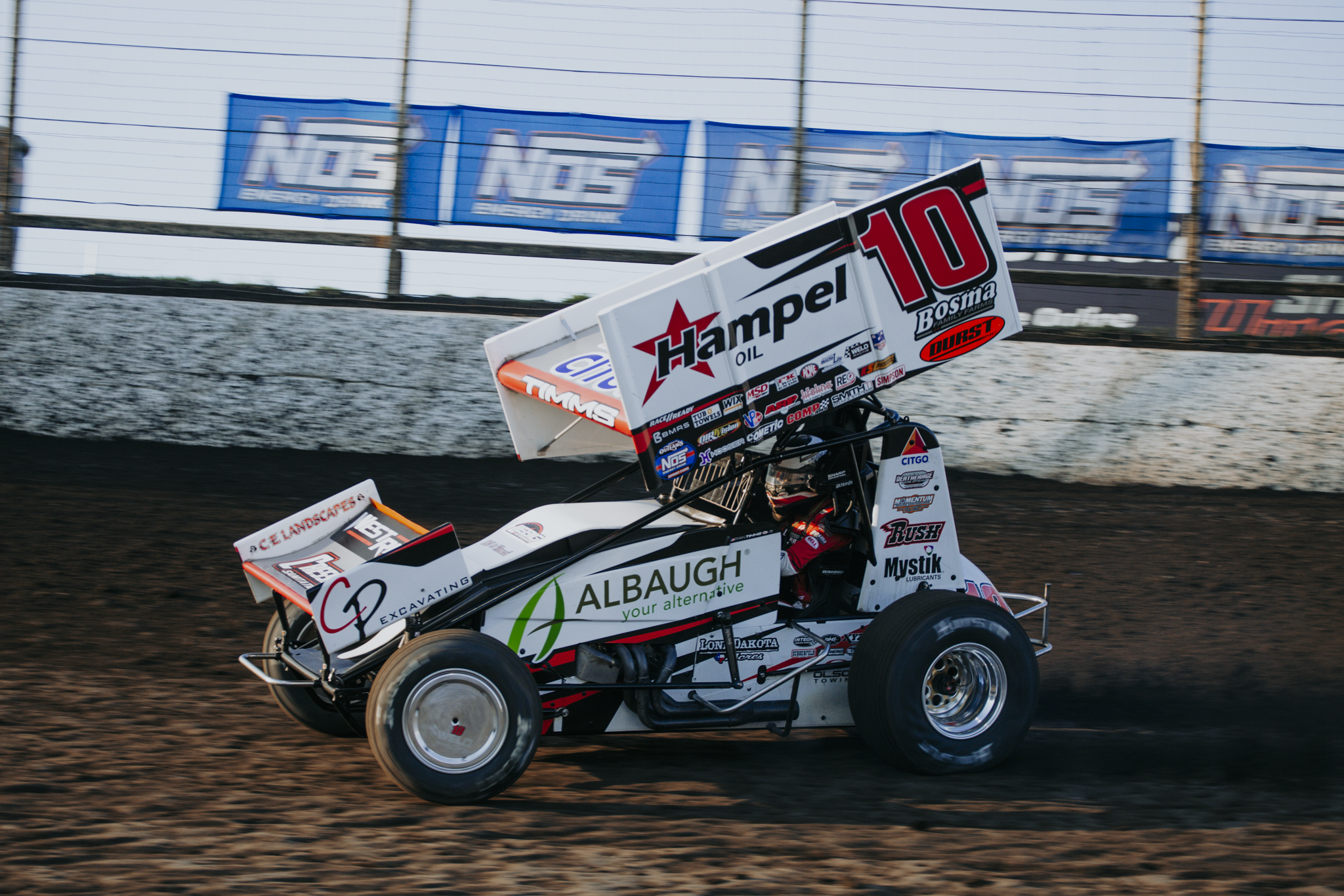 A sprint car on a dirt racetrack, featuring prominent sponsorship logos including Hamperl Oil and Albauh. The car is decorated in white, black, red, and green, with a large rear wing displaying the number 10. The background shows blurred advertisements and fencing, indicating motion and high speed.