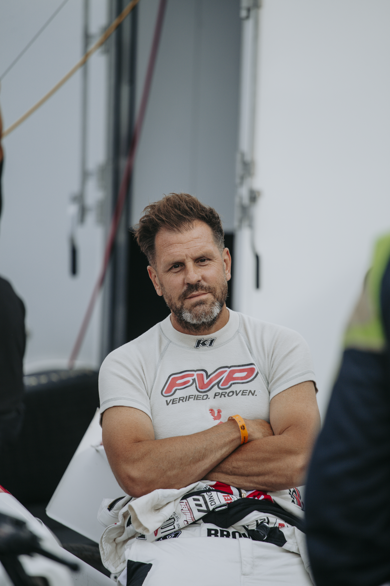 A man with a beard and short hair, wearing a white racing t-shirt, is standing with arms crossed, in an outdoor setting with a large equipment structure in the background. He's wearing an orange wristband and appears to be at a racing event.