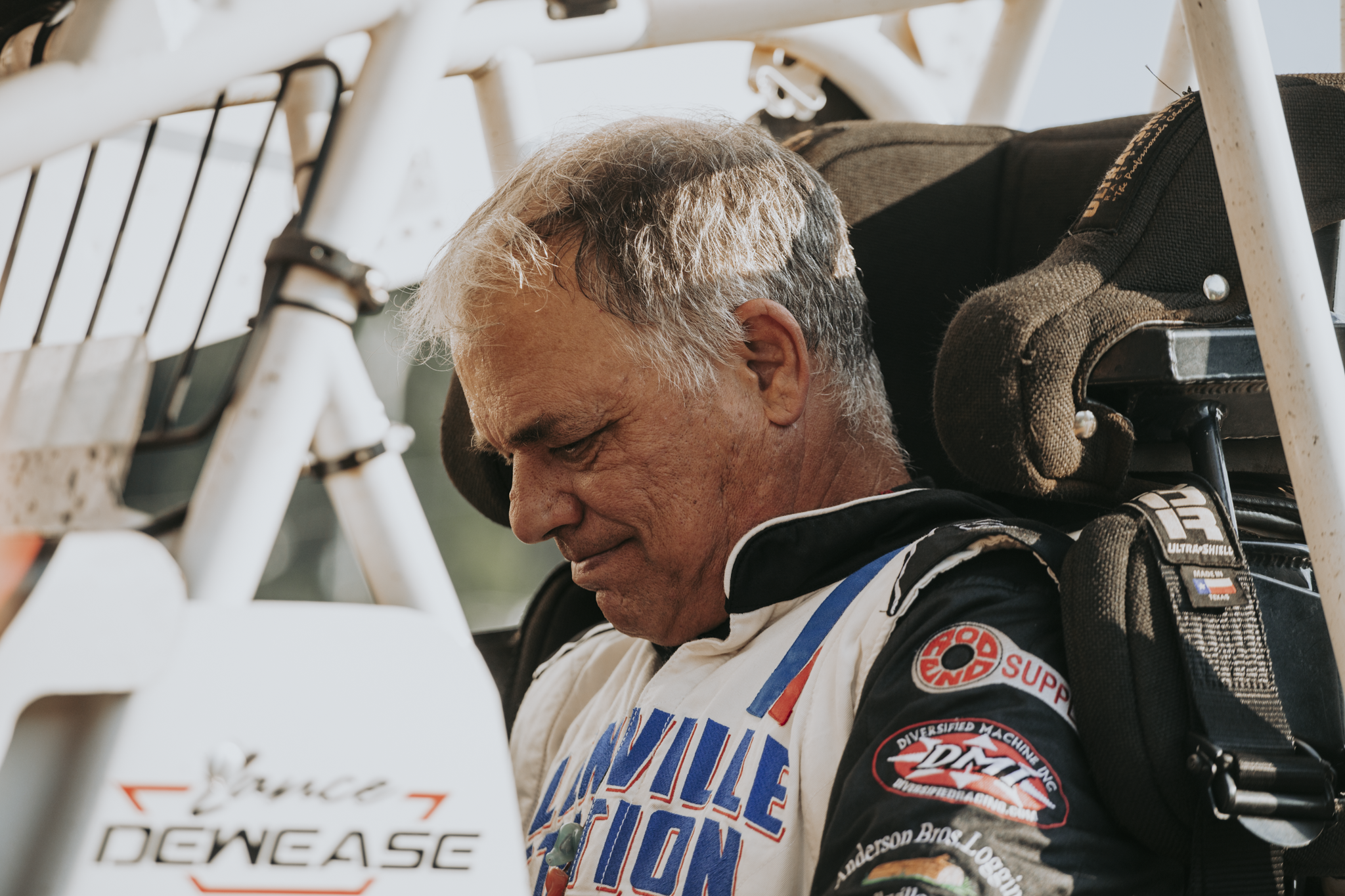 Close-up of an race car driver in racing suit, sitting in a racing vehicle with a white roll cage, looking down, at a racing event.
