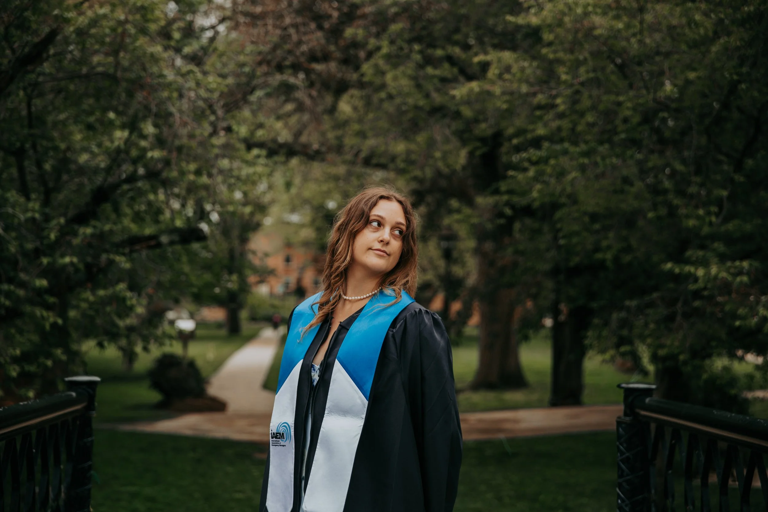 A young woman with wavy brown hair wearing a graduation gown and sash standing on a park bridge.