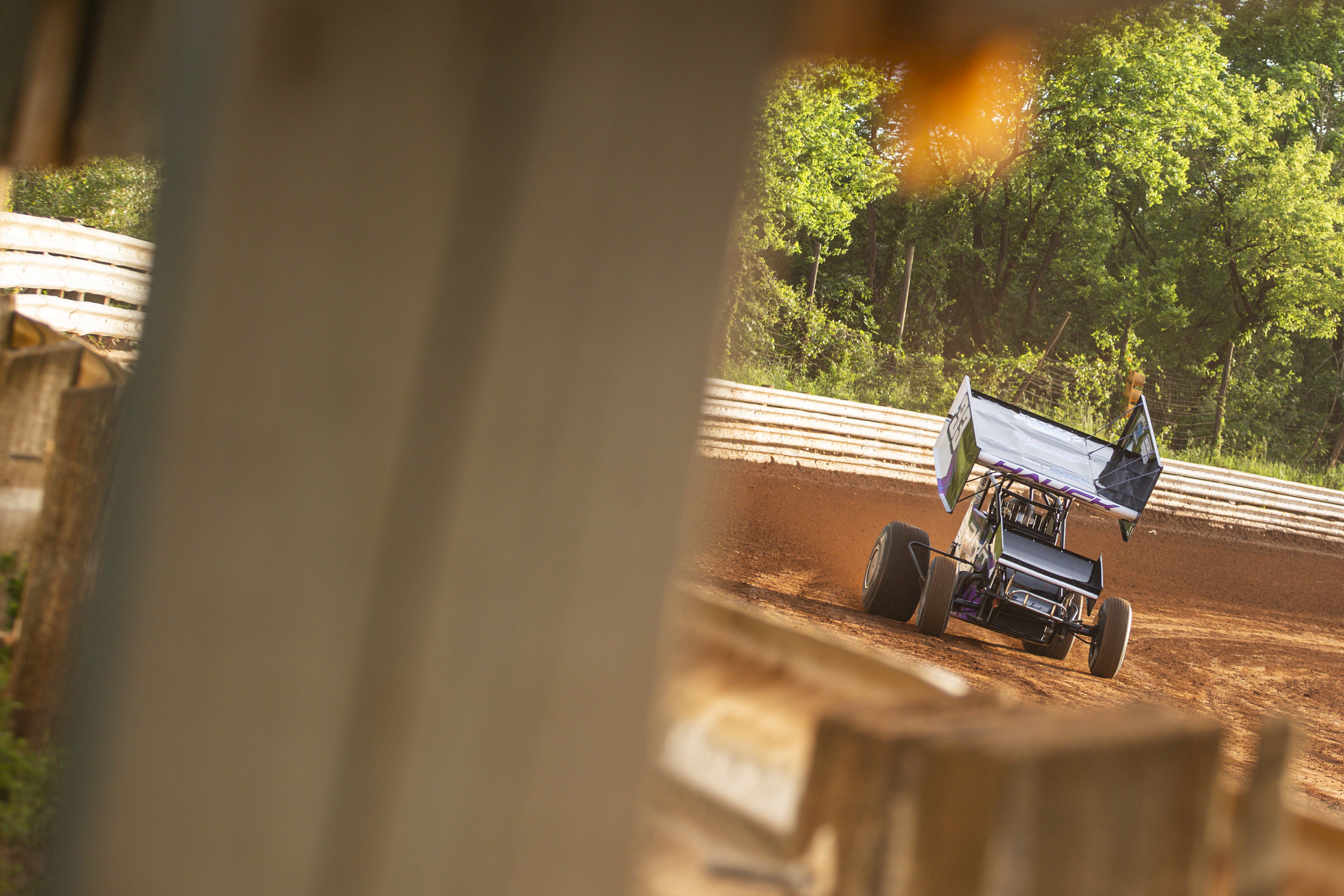 A dirt race track with a sprint car speeding around a corner, seen through a gap in a wooden fence. The background features green trees and a blue sky.