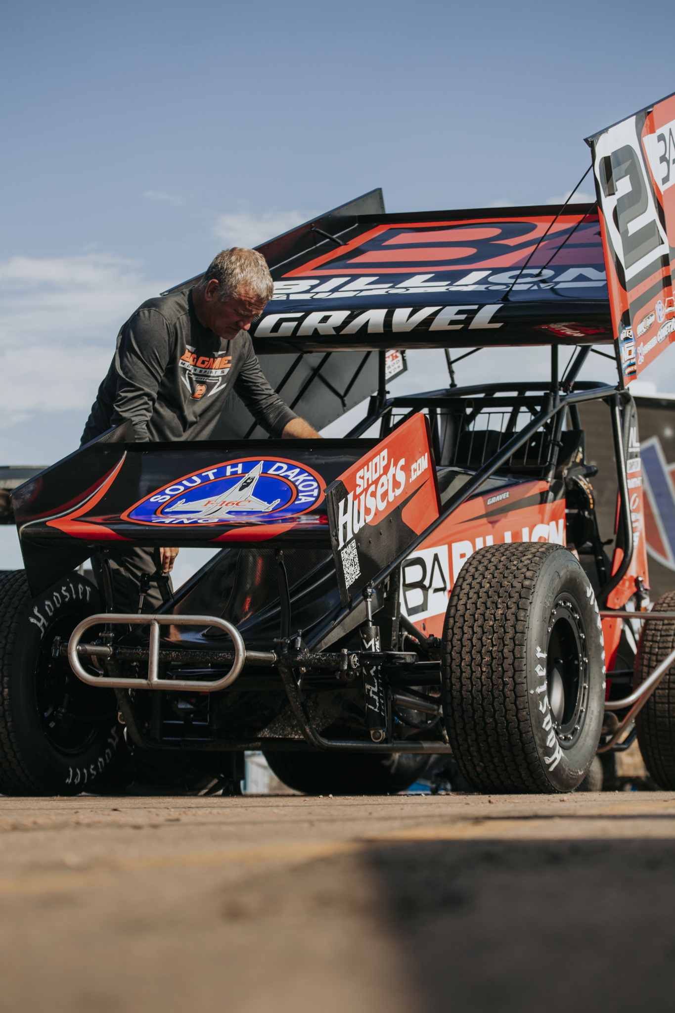 A man in a racing suit working on a black and orange sprint car with sponsorship logos, during a race event under a blue sky.