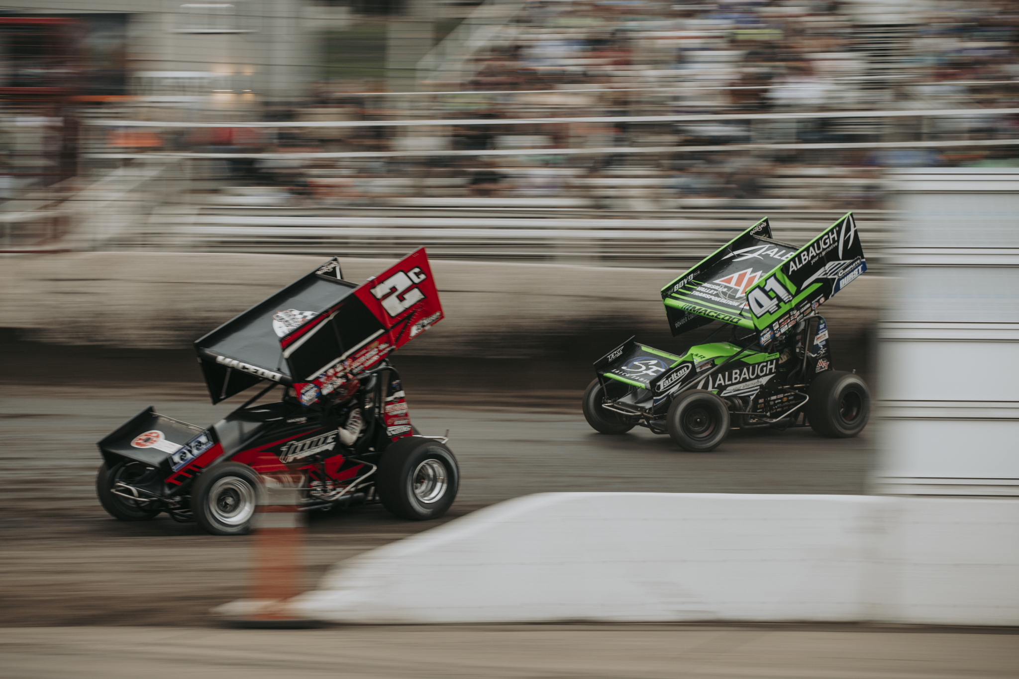Two sprint cars racing on a dirt track, with blurred crowd in the background, one green and black, and the other red and black.