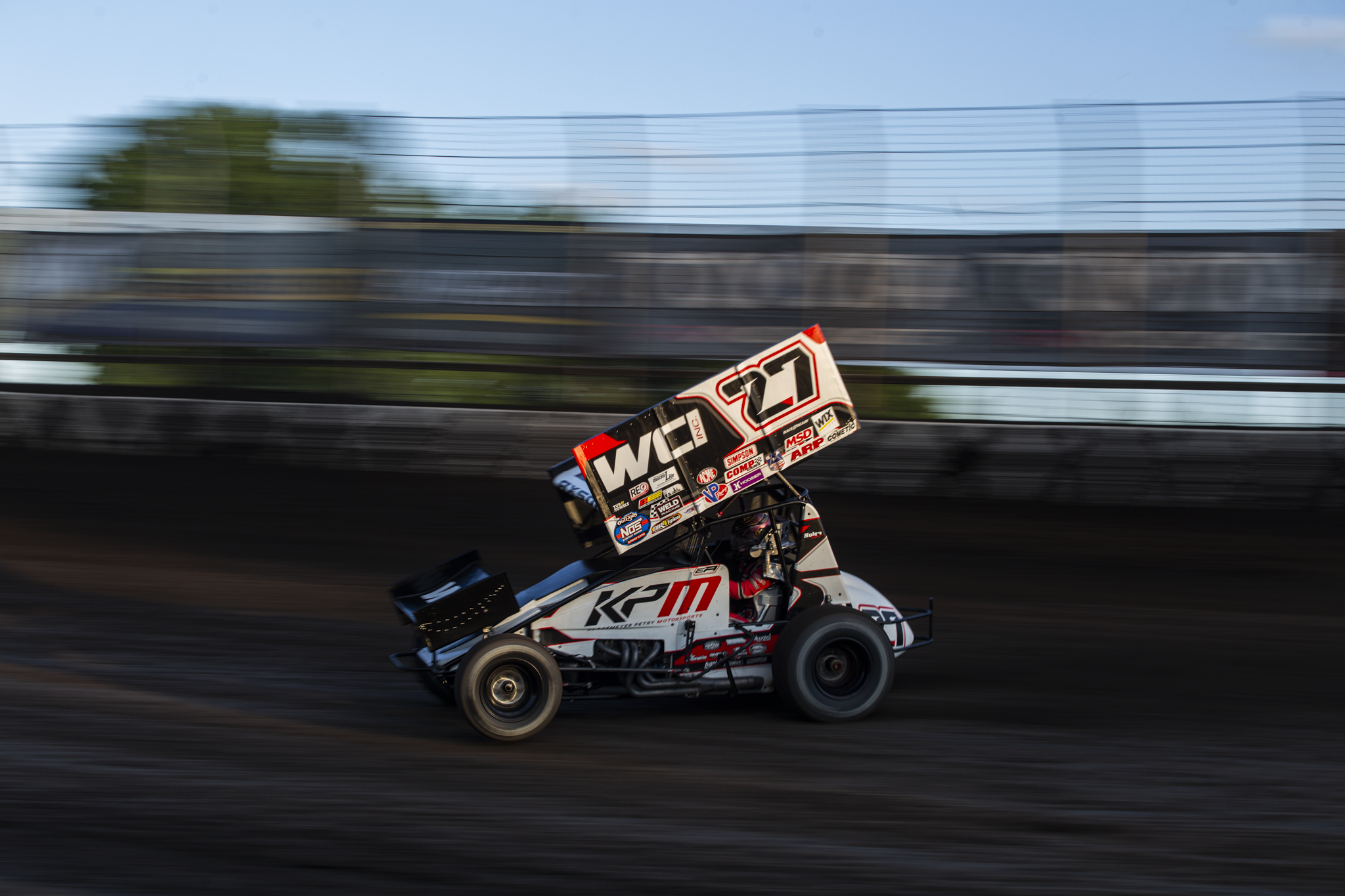 A sprint car racing on a dirt track at high speed, with motion blur in the background and a blurred background of trees and sky.