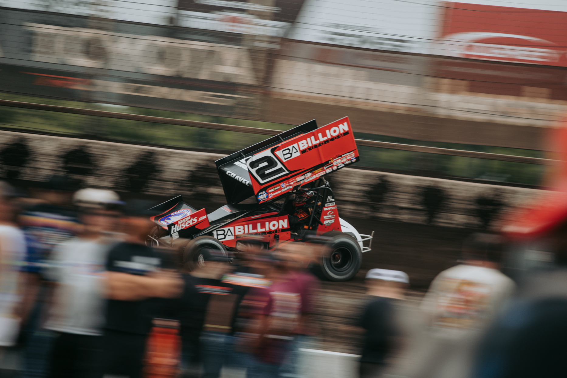 A sprint car racing on a dirt track with a blurred crowd of spectators in the foreground.
