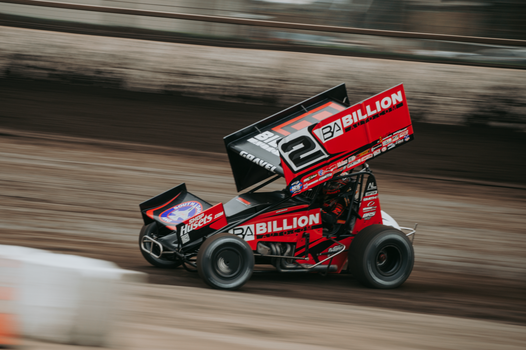 A sprint car racing on a dirt track with a large wing on top, featuring various sponsor logos including Billion Auto Group and Shop Huset's.